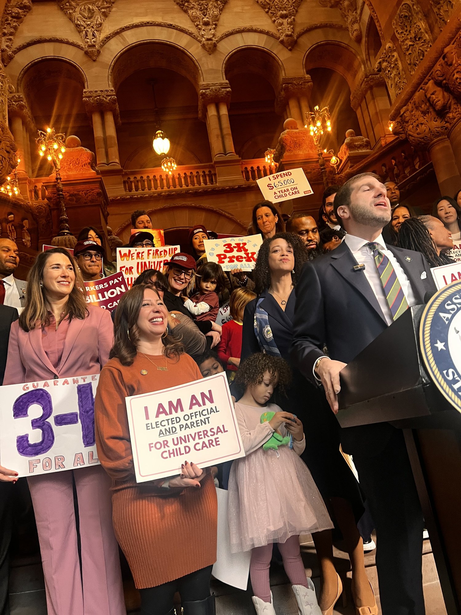 Advocates with 3-K signs and an elected official at a podium inside the New York State Capitol in Albany.