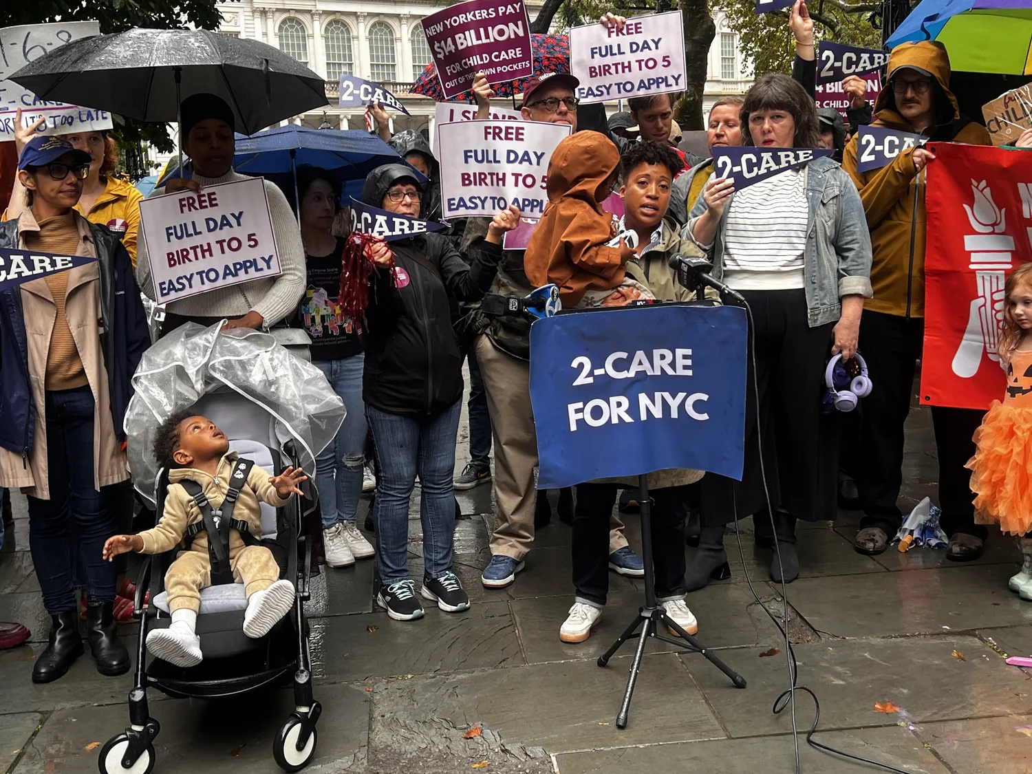 Parents with FREE FULL DAY BIRTH TO 5 signs behind a 2-Care for NYC podium at a New Yorkers United for Child Care rally.