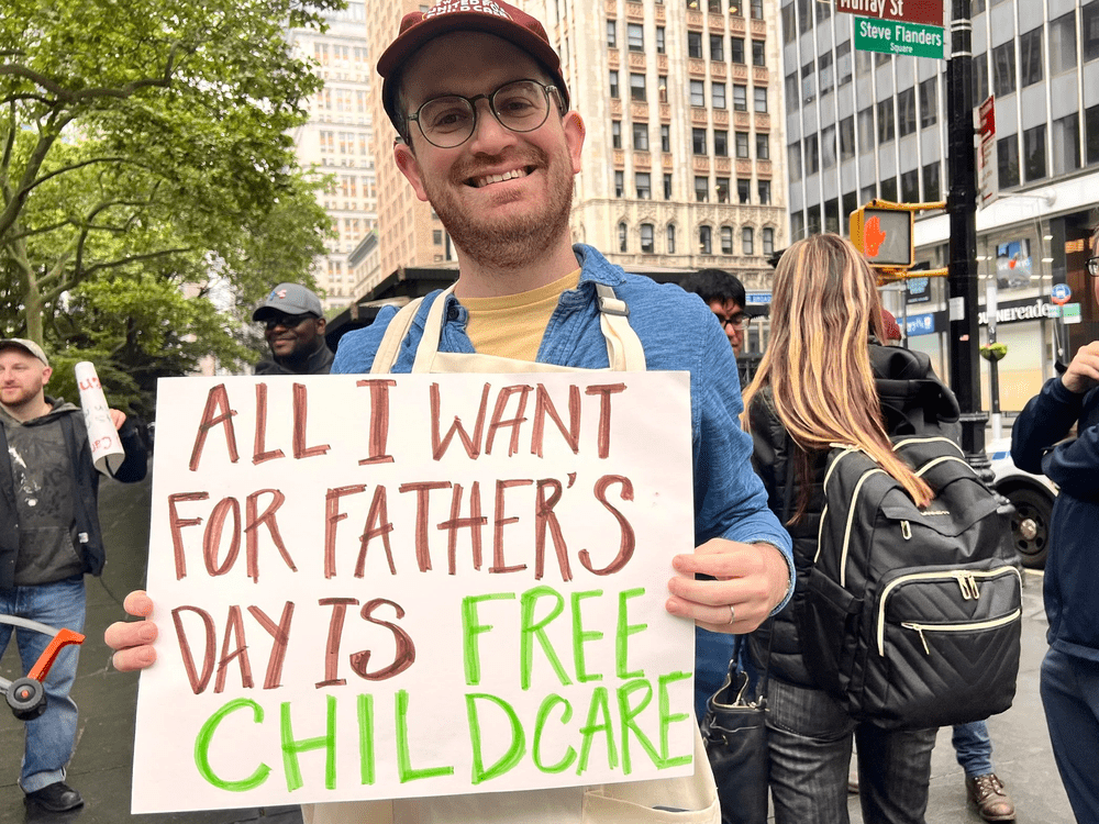A smiling dad in a cap holds a hand-painted sign reading “All I want for Father’s Day is free childcare” on a downtown street.