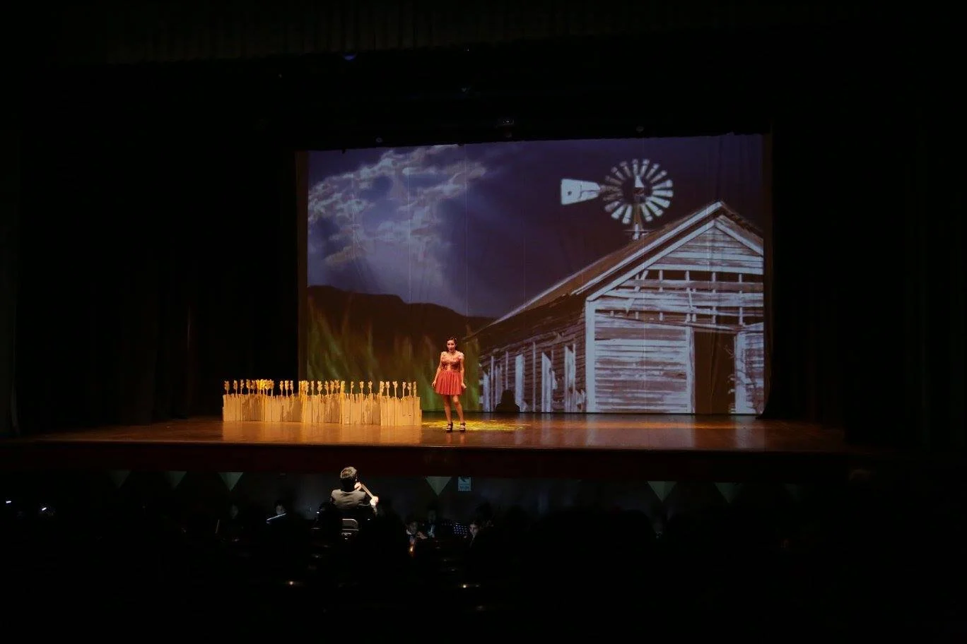 Stage scene showing a woman in a red dress standing in front of a backdrop with a barn, windmill, and sky scene. A person in the audience is taking a photo or video.