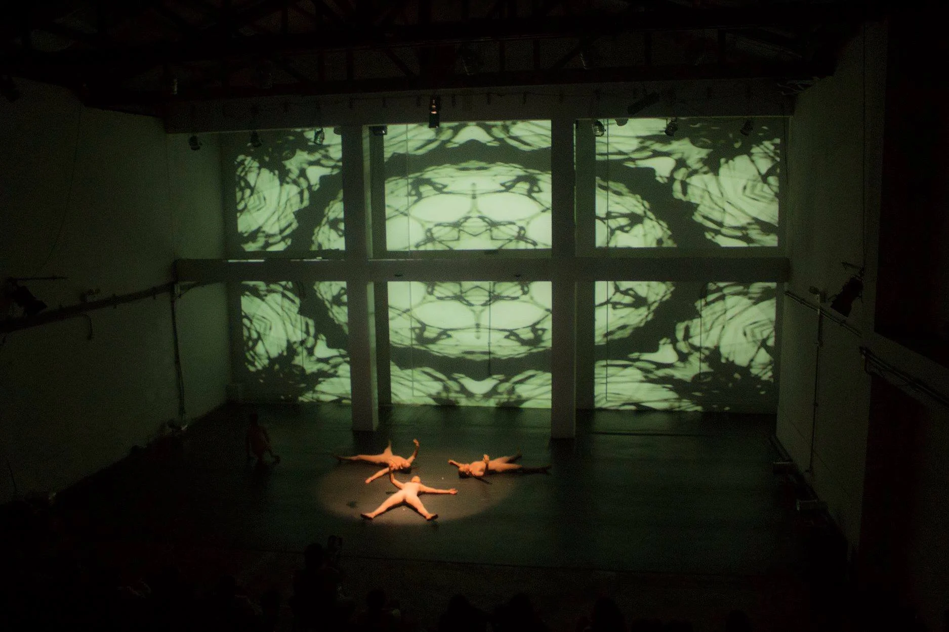 Dark theater stage with performers lying on the floor in a star shape, backlit by a large screen displaying abstract black and white patterns.
