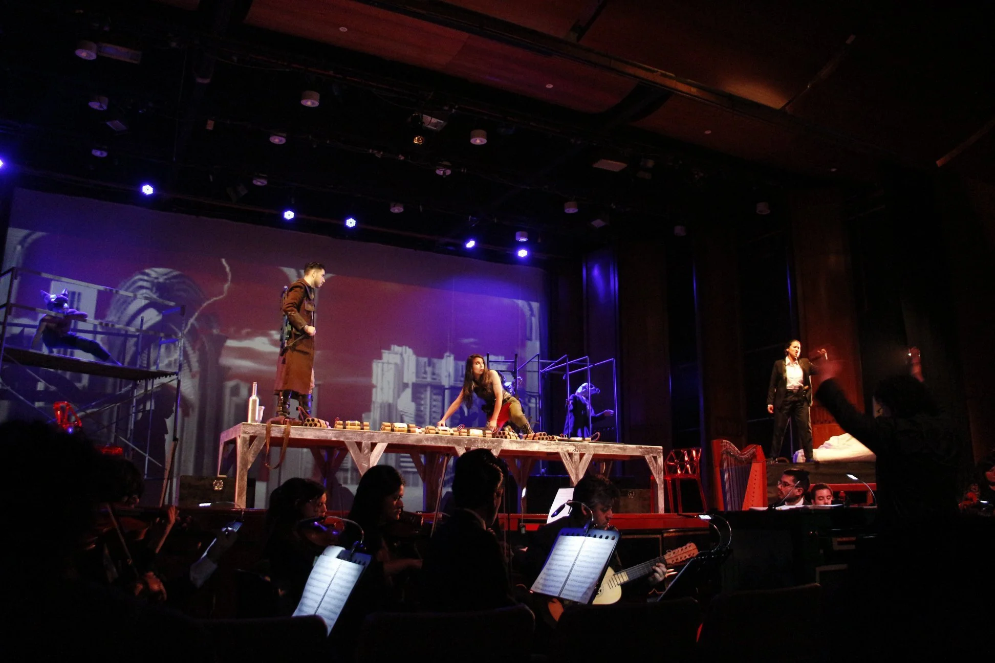 A stage with a theatrical performance featuring actors and musicians. One actor stands on a platform, another is seated on the stage floor, and others are positioned to the sides. The backdrop displays a cityscape image, and the stage is lit with blue and purple lights. Audience members are visible at the bottom, some holding scripts or musical scores.