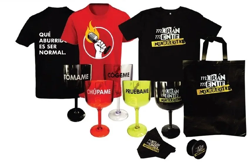 Collection of humorous-themed merchandise including T-shirts, glasses, a tote bag, a coaster, and a face mask. The items feature Spanish phrases and graphics, with some phrases like "Qué aburrido es ser normal," and words such as "Tómame," "Cógeme," "Chúpame," and "Pruébame" on the glasses. The shirts display phrases about morbid or dark humor with graphic designs, including a burning microphone and text overlay.