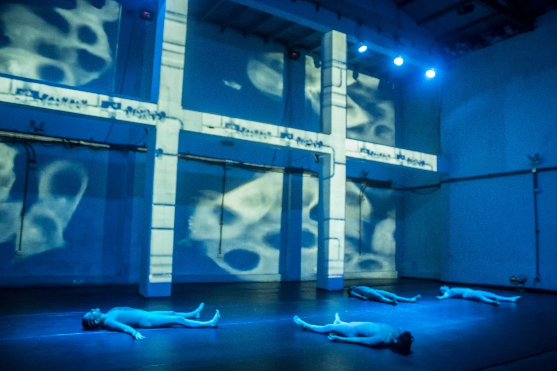 Dimly lit stage with four people lying flat on their backs, spread out apart, in front of a large set piece resembling building walls with clouds projected on it. Blue stage lighting casts shadows, creating an eerie atmosphere.