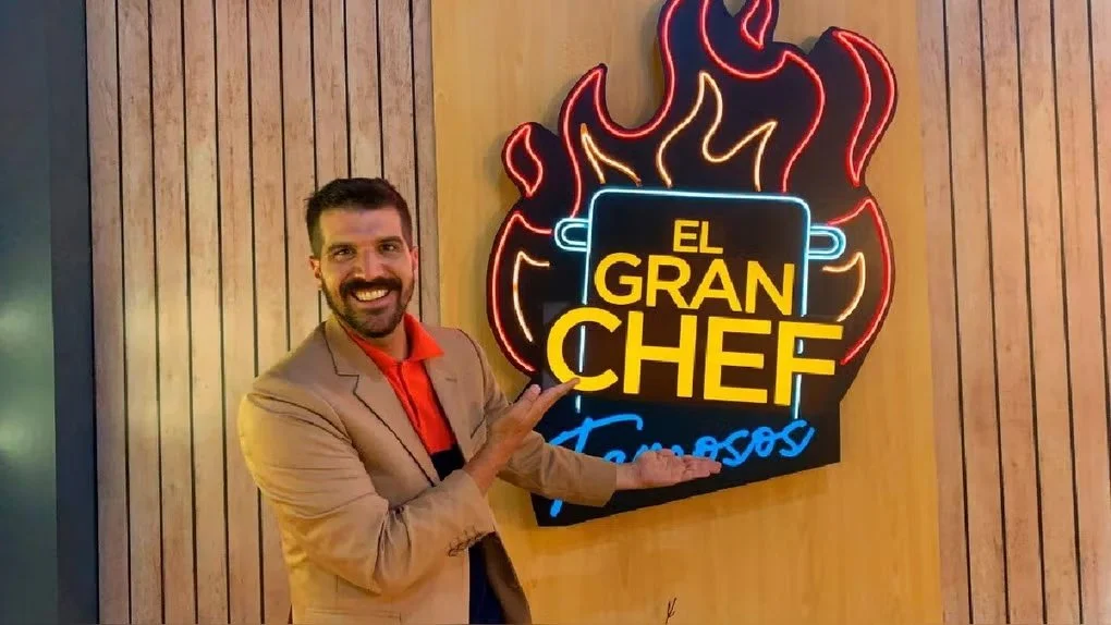 A man in a beige blazer and red shirt pointing at a neon sign that says 'EL GRAN CHEF' with flames in the background, mounted on a wooden wall.