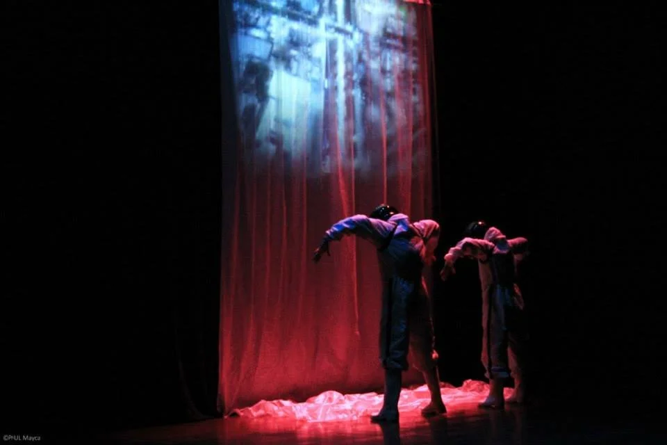 Two dancers on stage under a red spotlight, bent forward in performance, with a red curtain and projected imagery behind them.