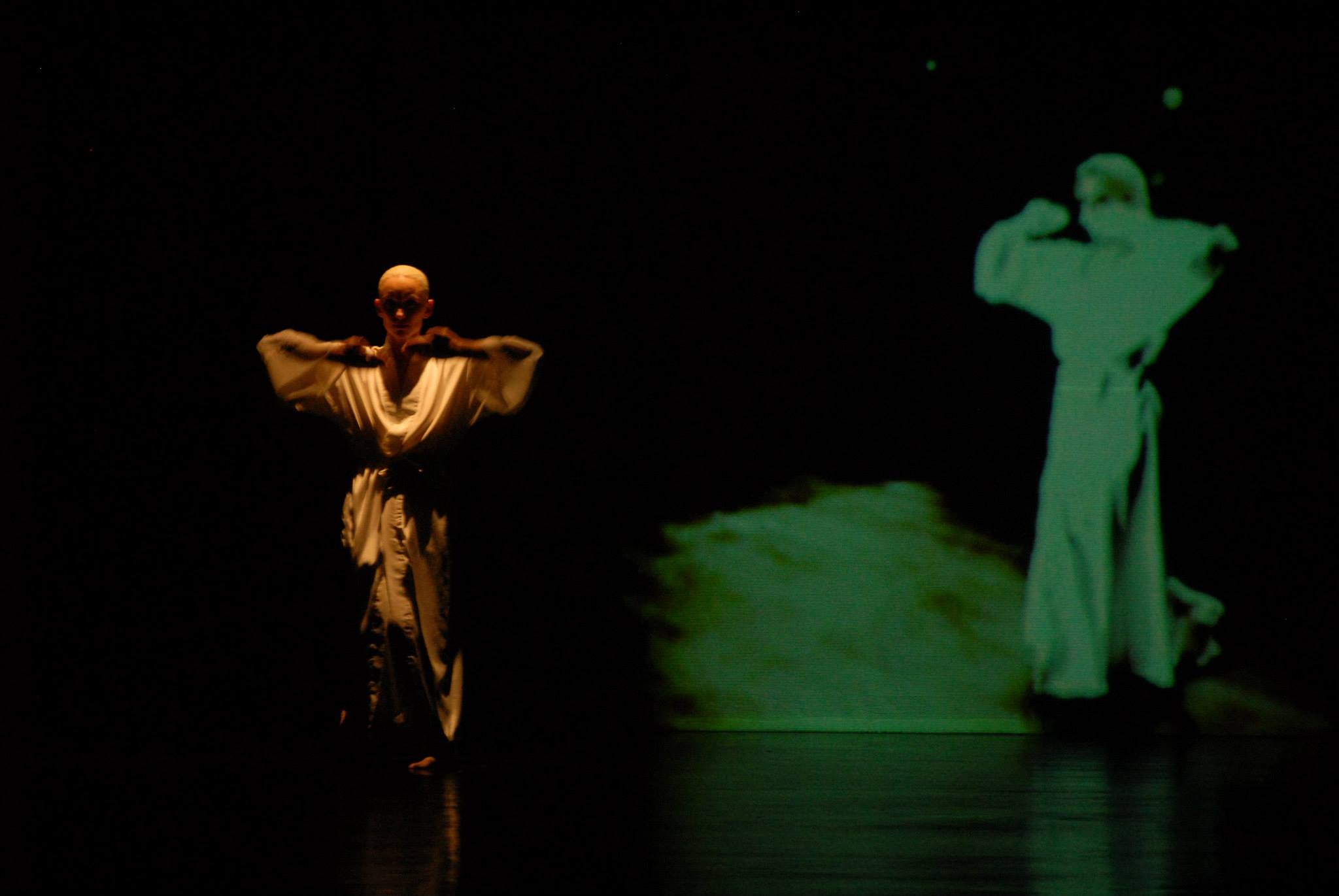 A performer in a beige costume strikes a pose on stage with a dark background, while a green-tinted statue of a woman with a dog appears to be part of a projection or display to the right.
