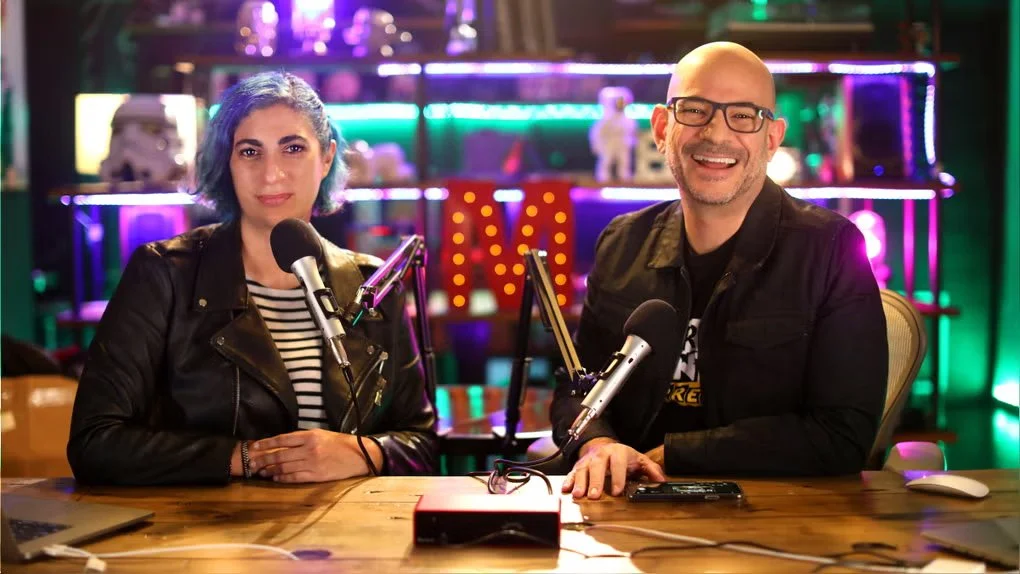 Two people sitting at a podcast recording studio with microphones, smiling at the camera, colorful neon lights, and a decorated background.