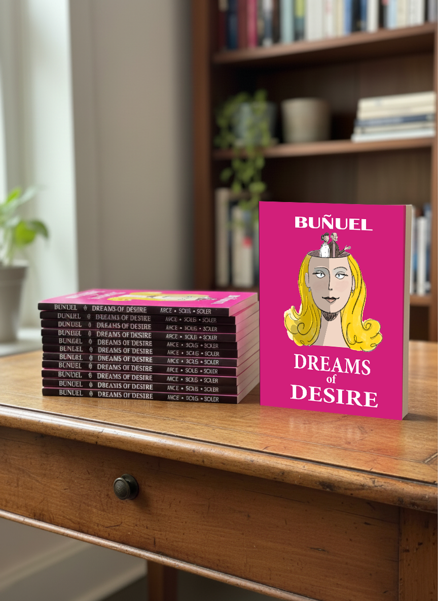 A pink book titled 'Dreams of Desire' by Bunuel standing upright on a wooden table, with multiple copies stacked next to it. In the background, a bookshelf with various books and a potted plant is visible.