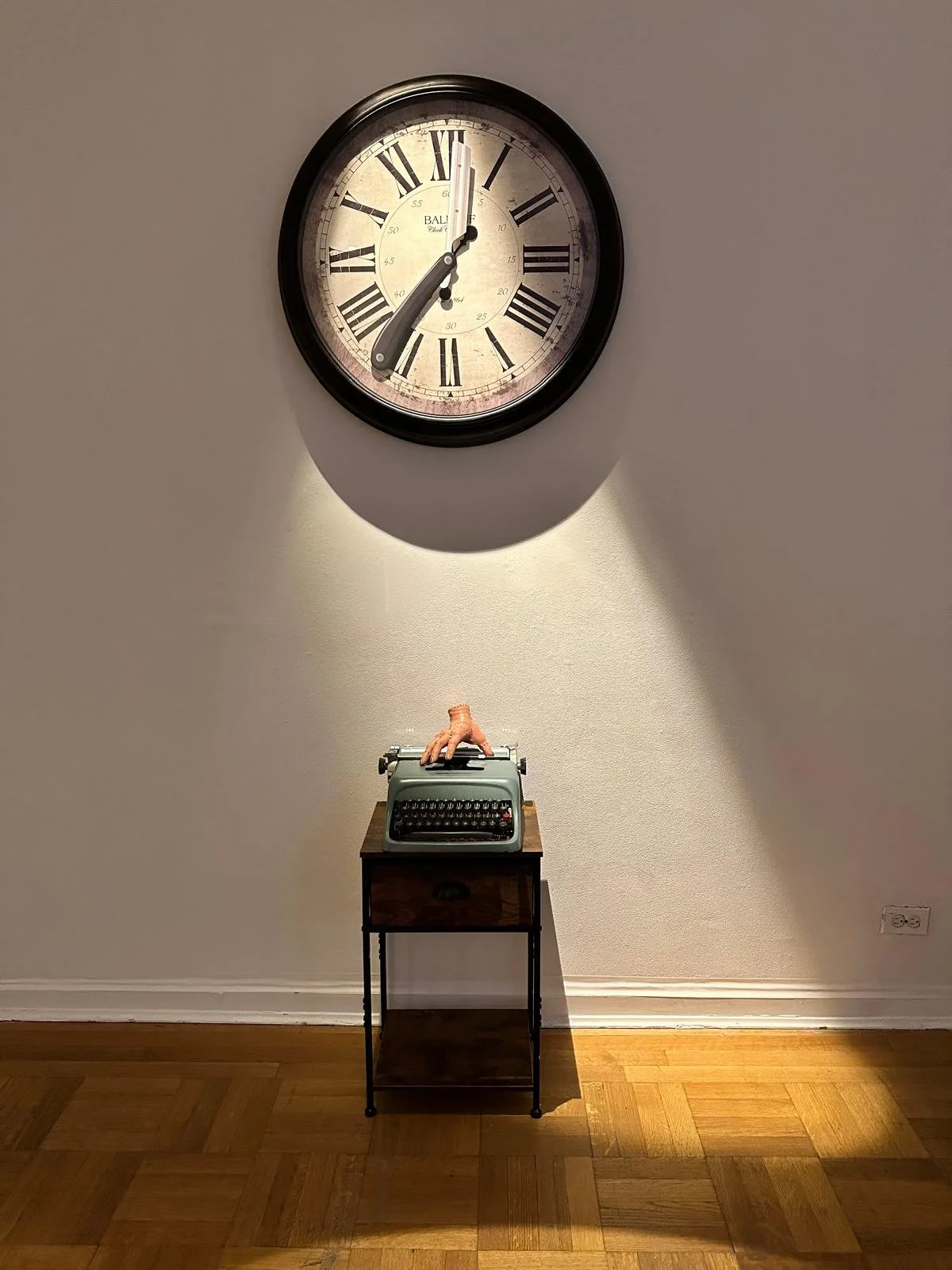 A large round clock hanging on a wall showing 12:00. Below the clock is a vintage typewriter placed on a small wooden table, with a rubber hand on top. The wall is plain and painted in a neutral color.