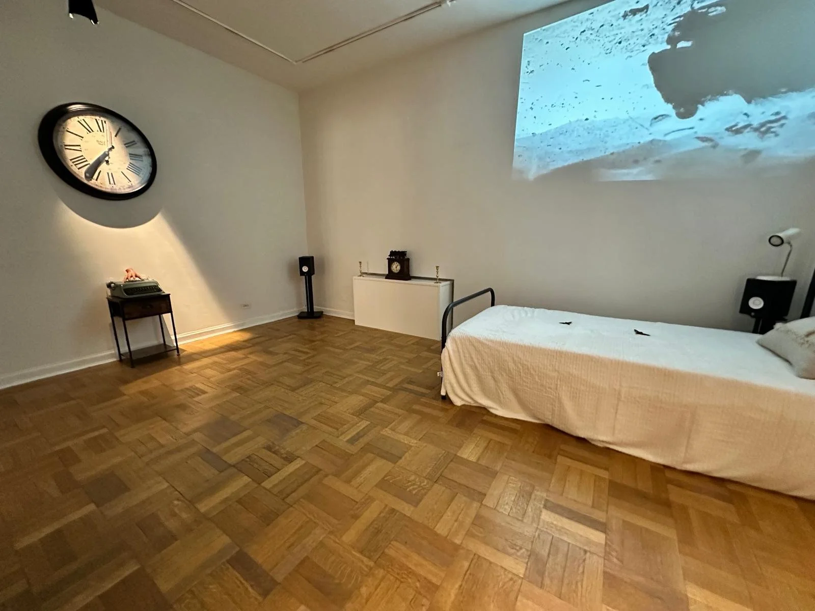 A minimalist bedroom with a wooden floor, a bed with white bedding, a wall-mounted clock, a small side table with a vintage telephone, and a wall projection of a beach scene.