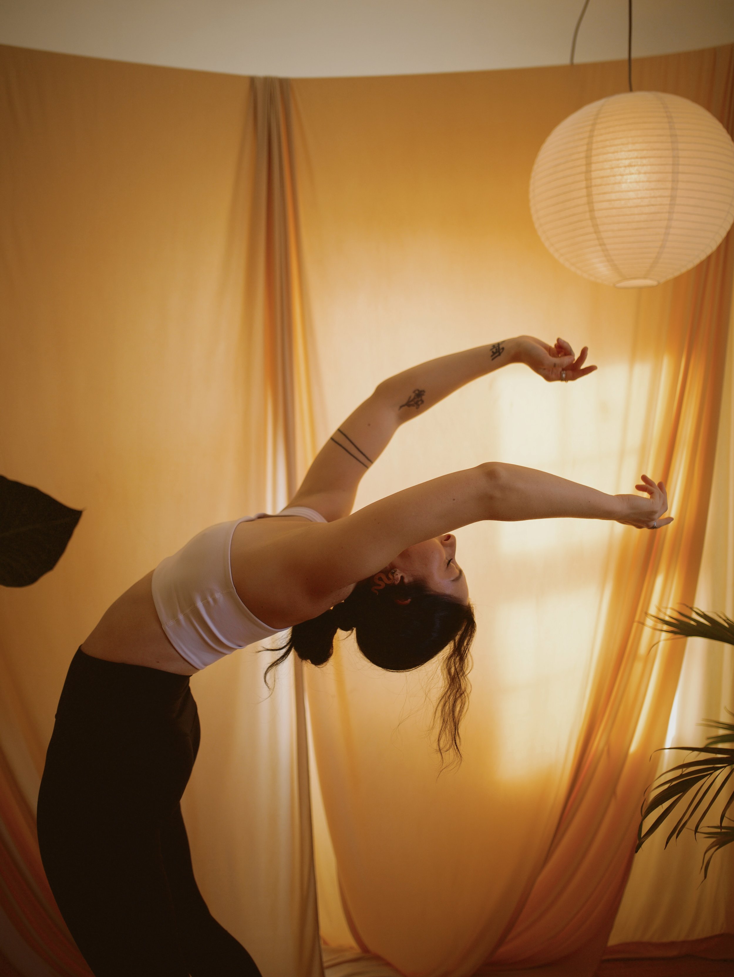Black and white photo of two women practicing yoga or meditation indoors, with one sitting cross-legged and hands in prayer position, and the other performing a stretching pose with one arm extended and the head resting on the knee.