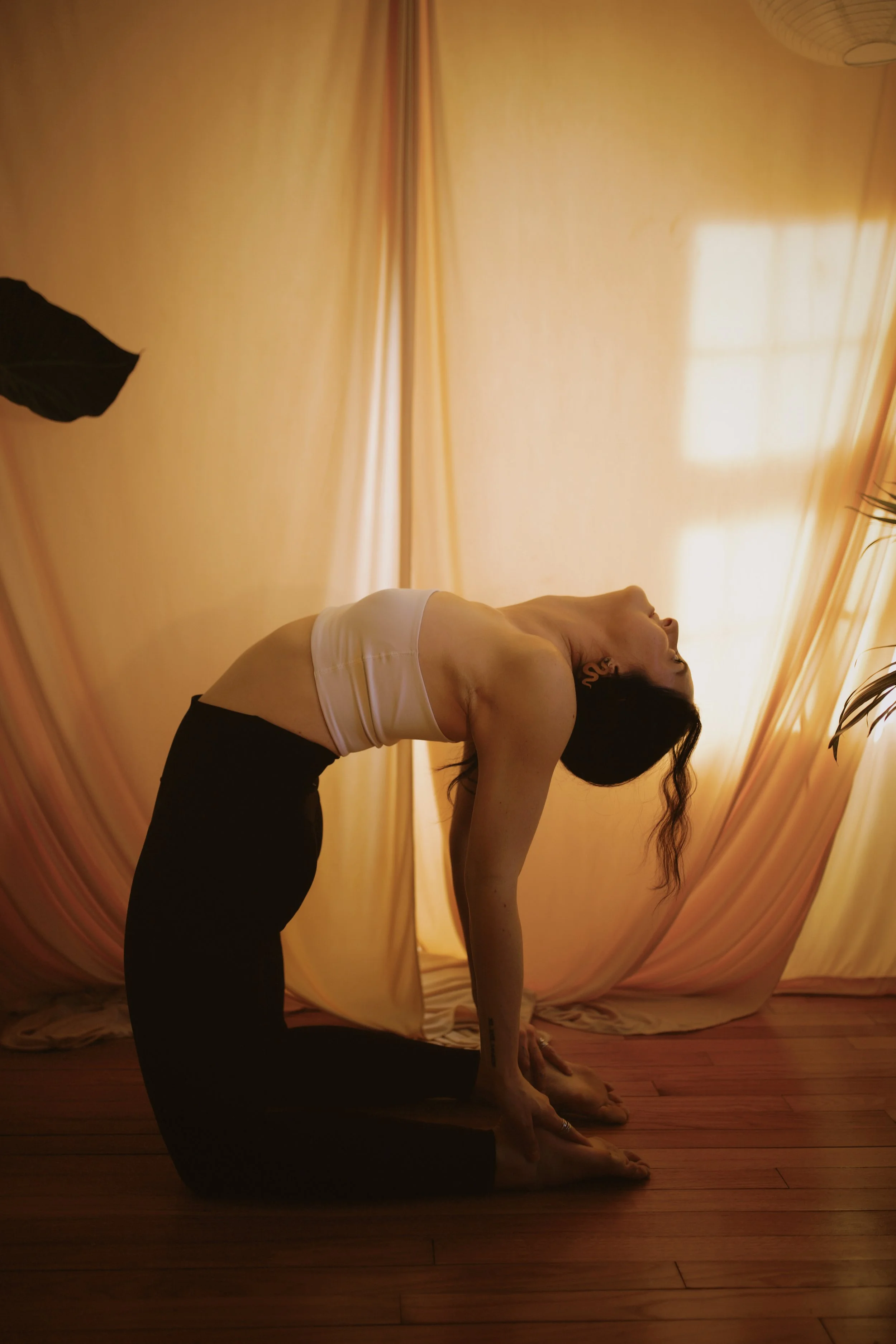 A woman sitting cross-legged on a wooden floor, practicing yoga or meditation, with a neutral expression, against a plain wall.