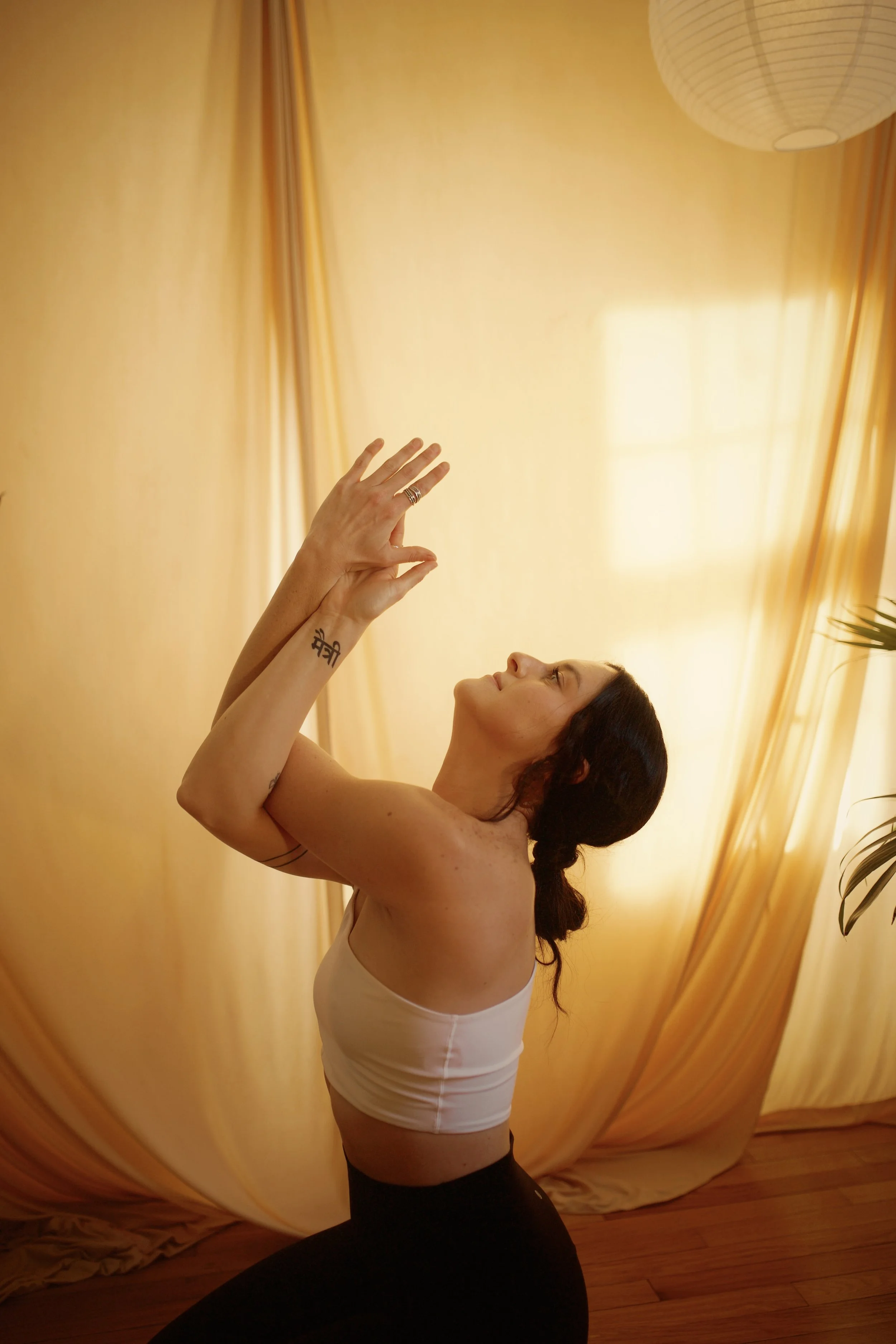 A woman sitting cross-legged on a wooden floor, practicing yoga or meditation, with a neutral expression, against a plain wall.