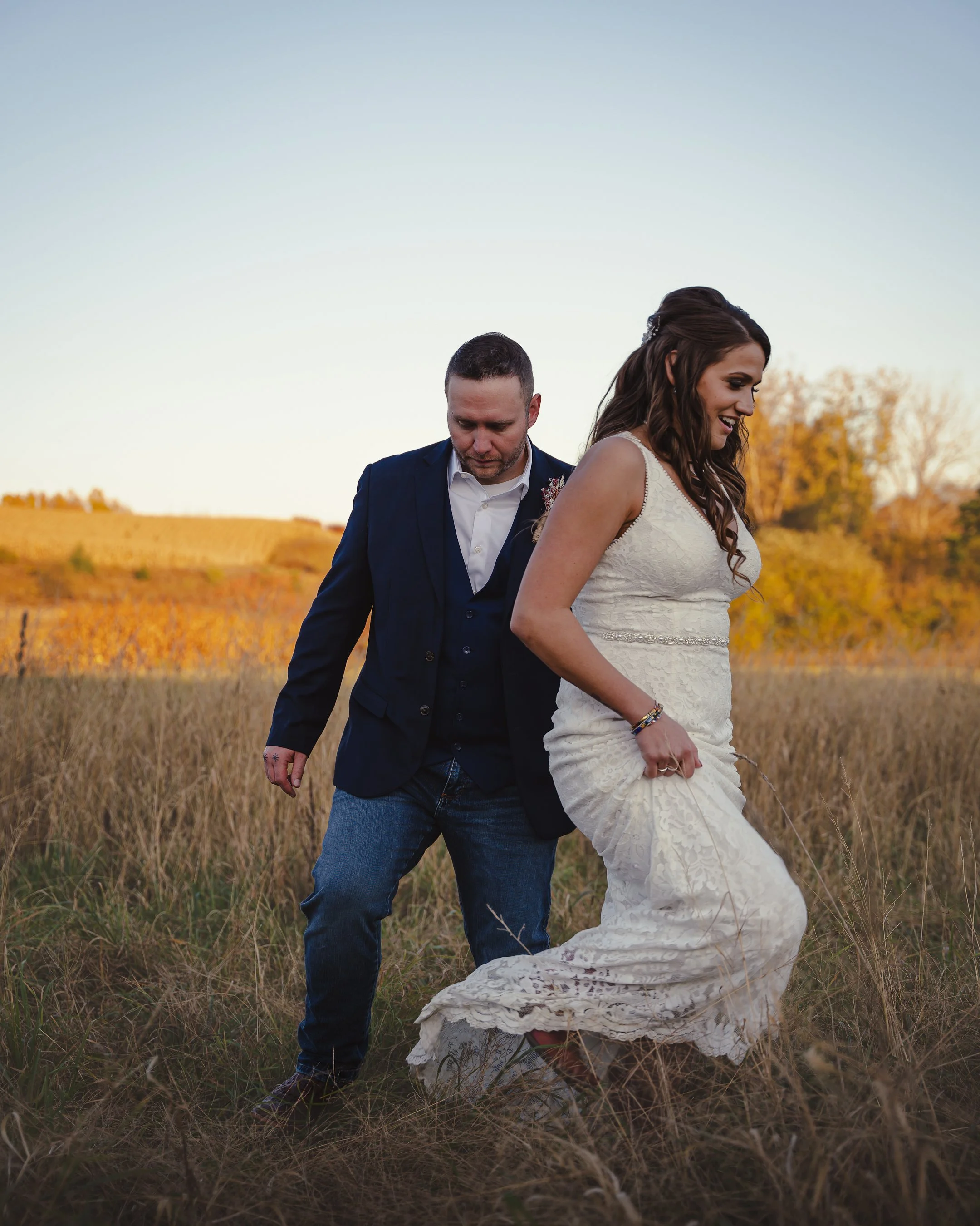 A couple in wedding attire, a man in a dark suit and a woman in a white dress, standing in a field of tall grass during sunset.