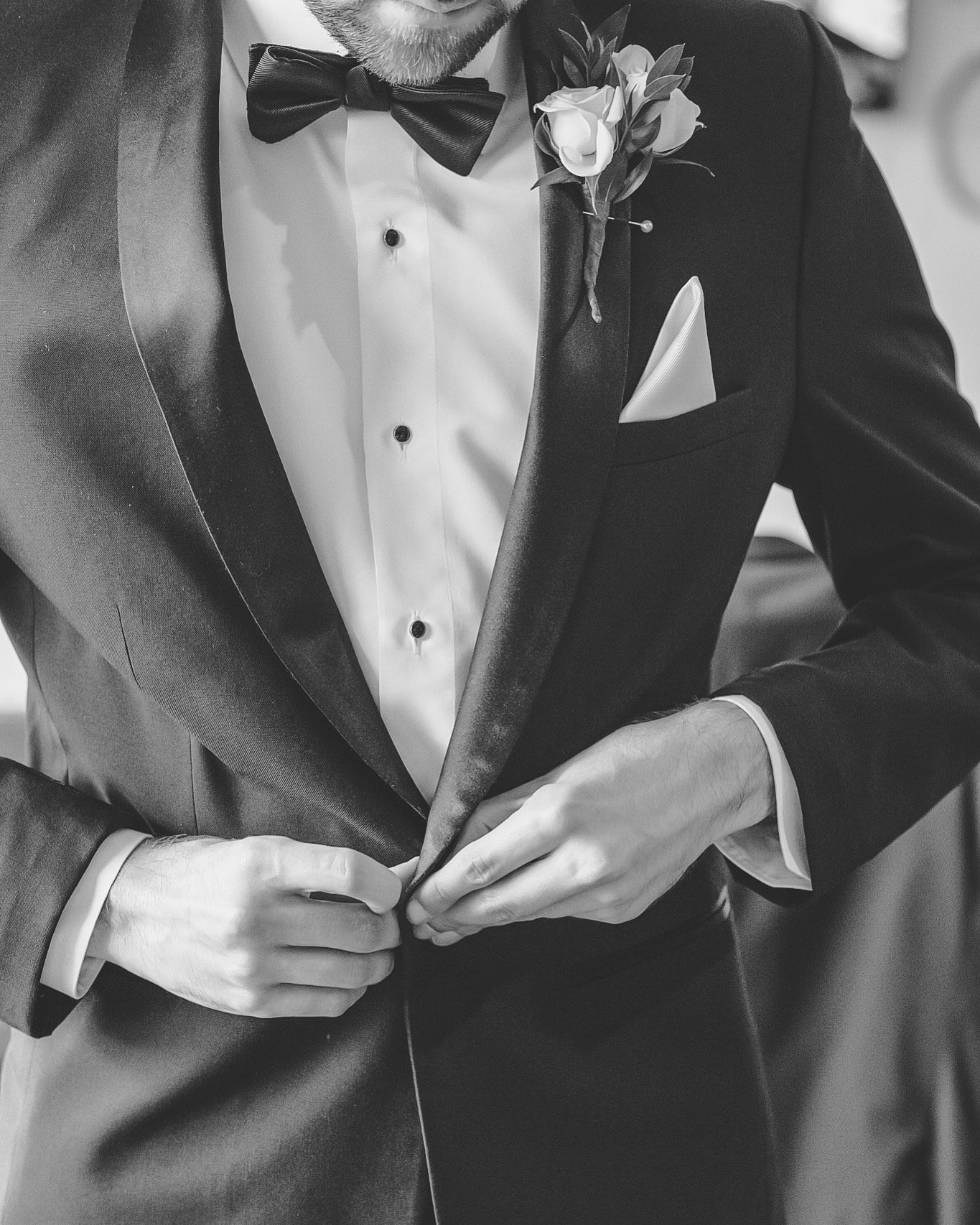 Close-up of a man in formal black tuxedo with a white shirt, black bowtie, and boutonniere, buttoning his jacket.
