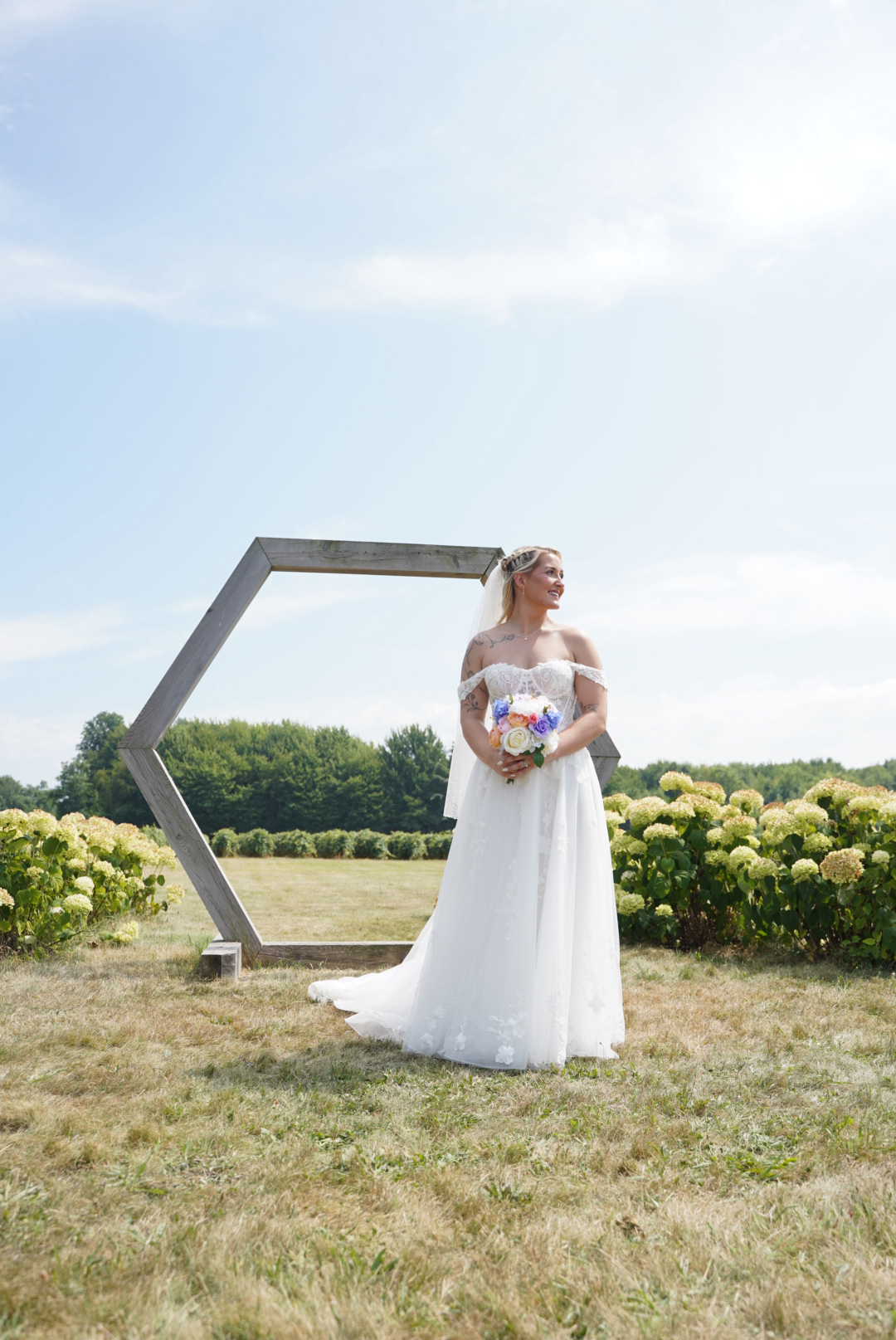 A bride in a white wedding dress holding a bouquet of flowers standing outdoors near a geometric wooden structure surrounded by green shrubs and trees