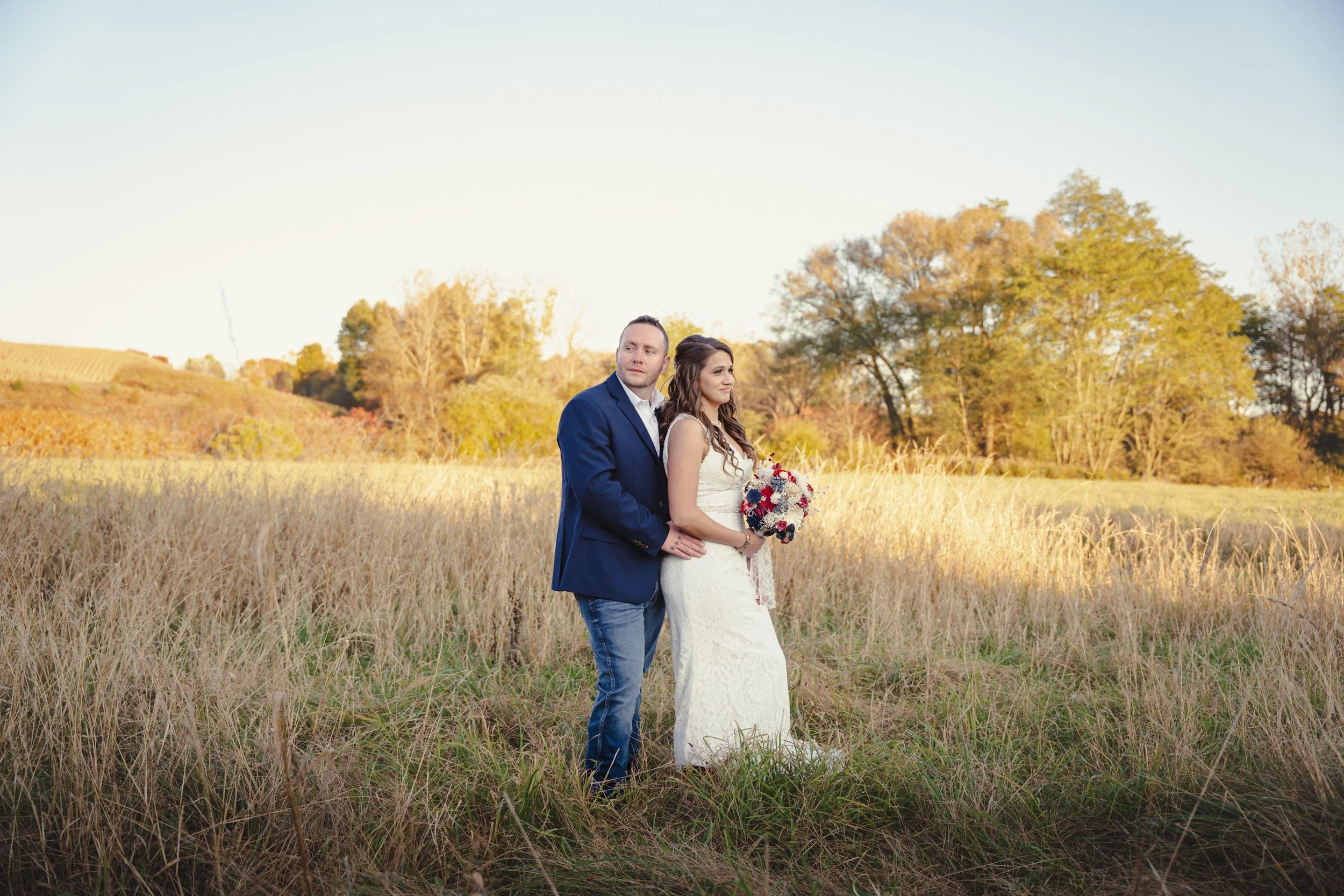 A bride and groom standing in a field of tall grass during sunset, with trees and a hill in the background, celebrating their wedding.