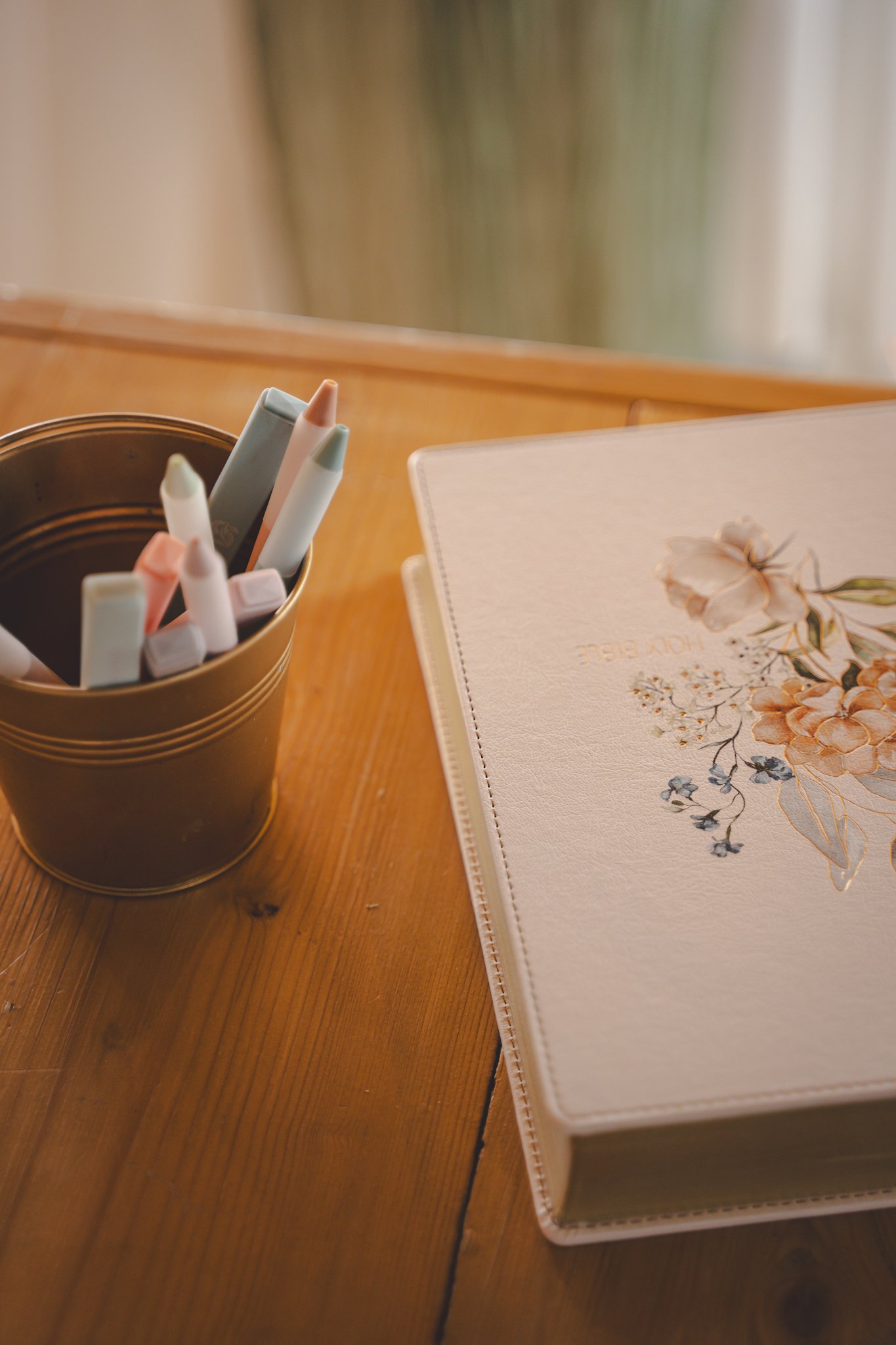 A floral planner and a pen holder with pastel-colored pens on a wooden desk.