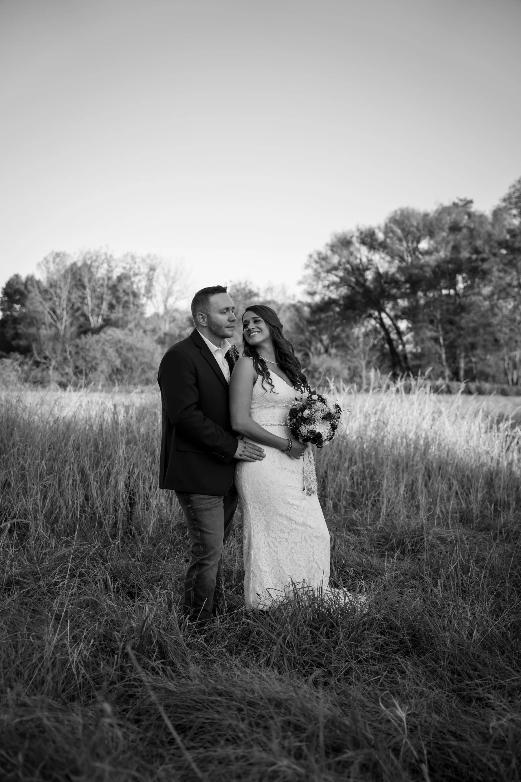 Black and white photo of a bride and groom standing in a field with trees in the background, the bride holding a bouquet, both smiling