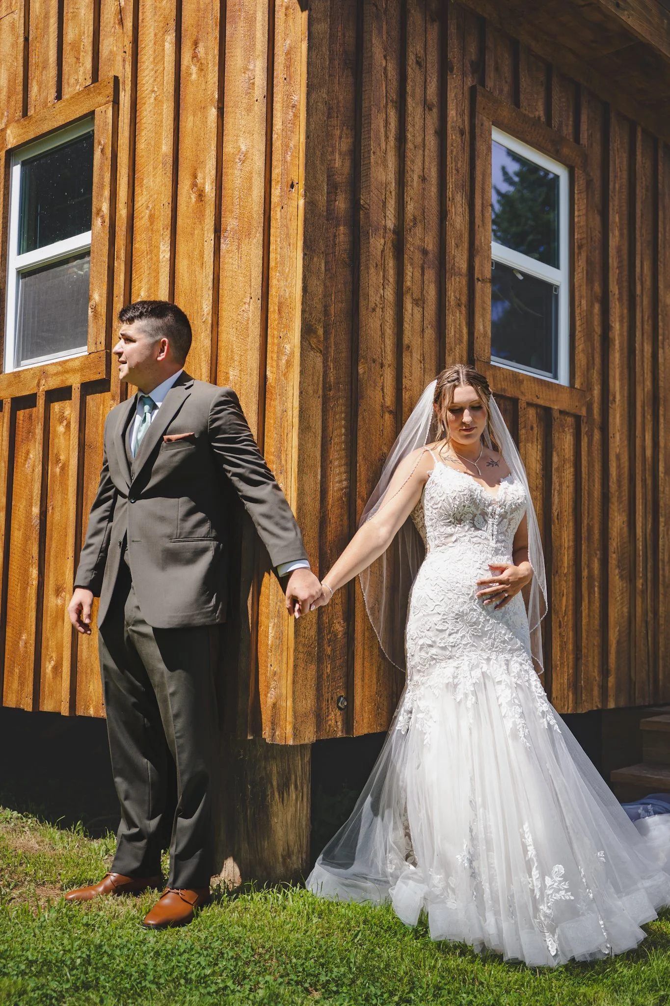 A bride in a white wedding dress and a groom in a gray suit holding hands beside a wooden building.