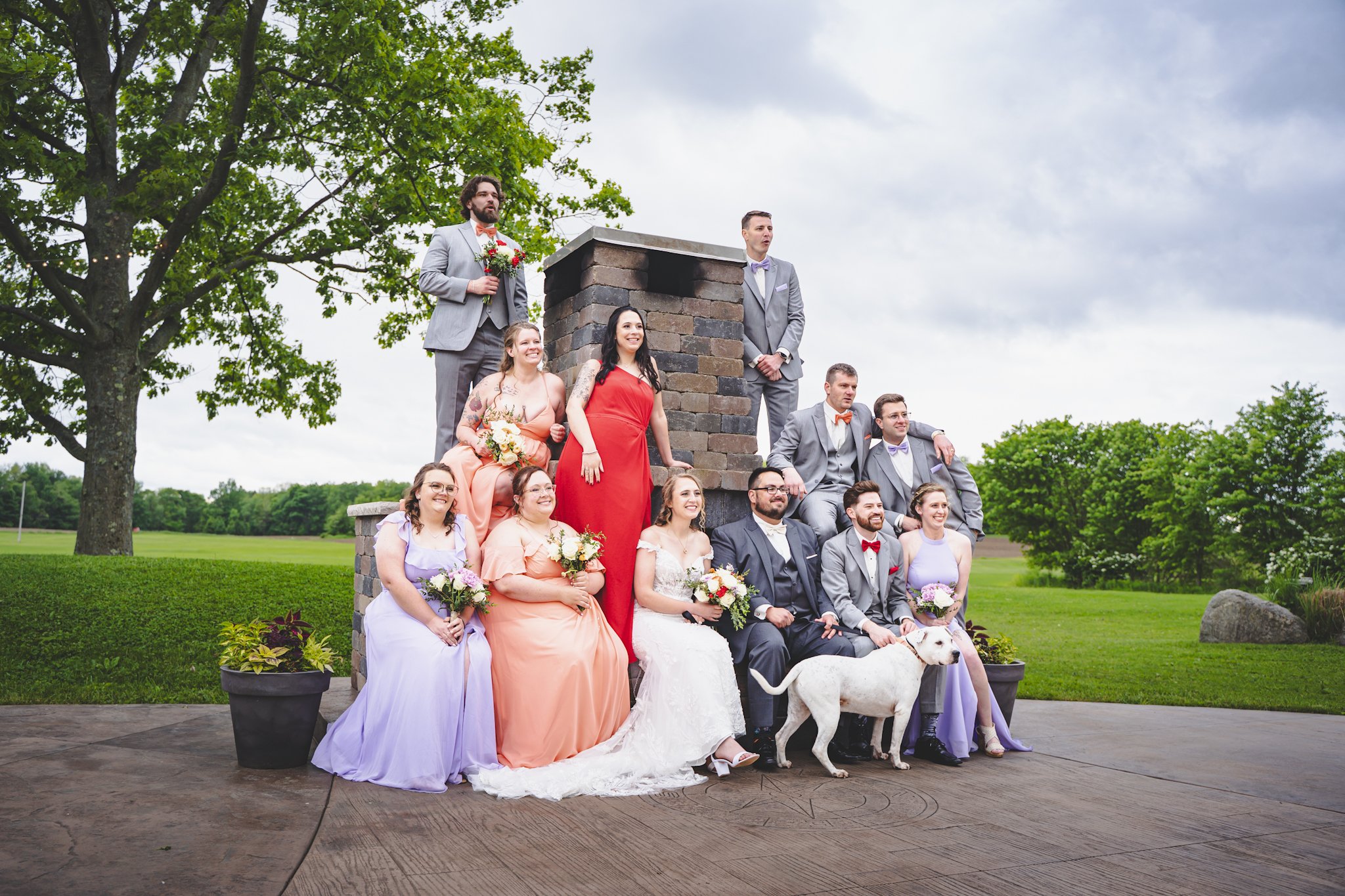 Group of people dressed in formal wedding attire sitting and standing around a brick structure outdoors on a cloudy day, with green trees and grass in the background.