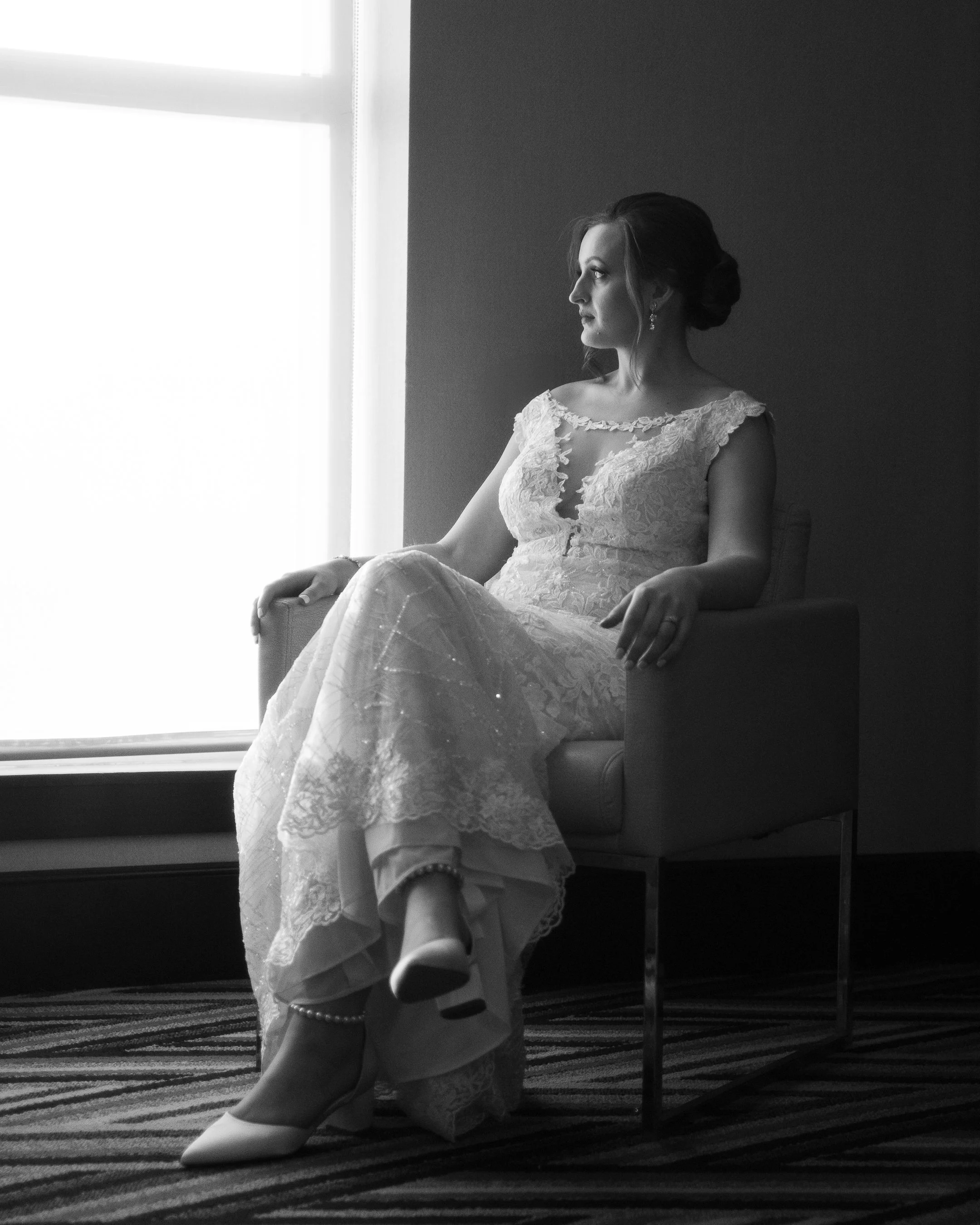 Black-and-white photo of a woman in a lace wedding dress sitting on a chair, looking out the window.