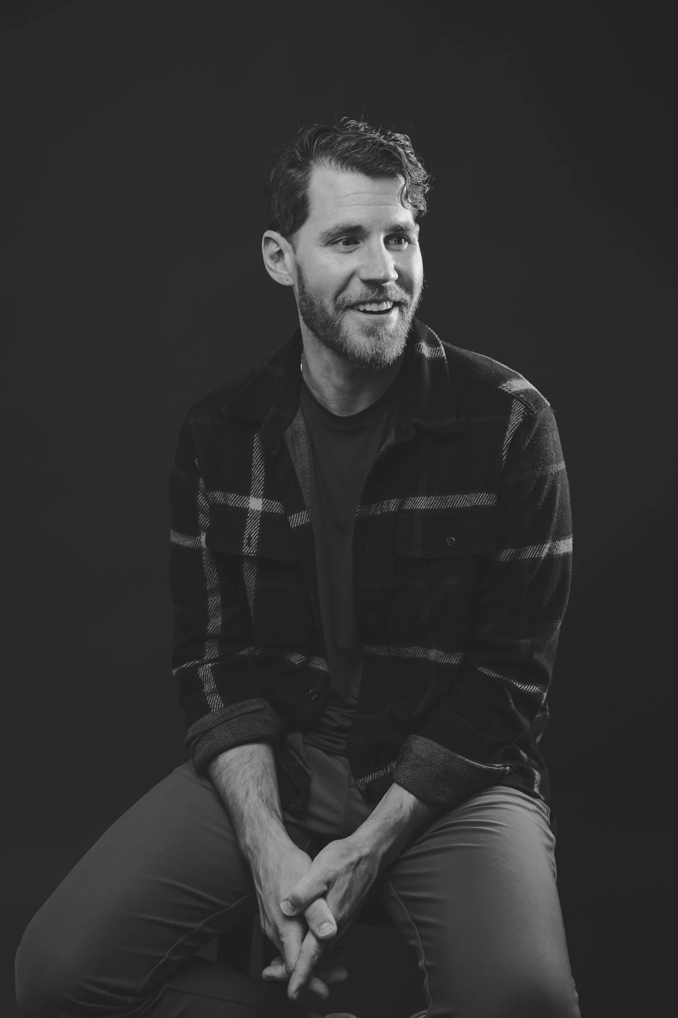 Black and white portrait of a smiling man with a beard, sitting cross-legged against a dark background, wearing a plaid shirt and casual pants.