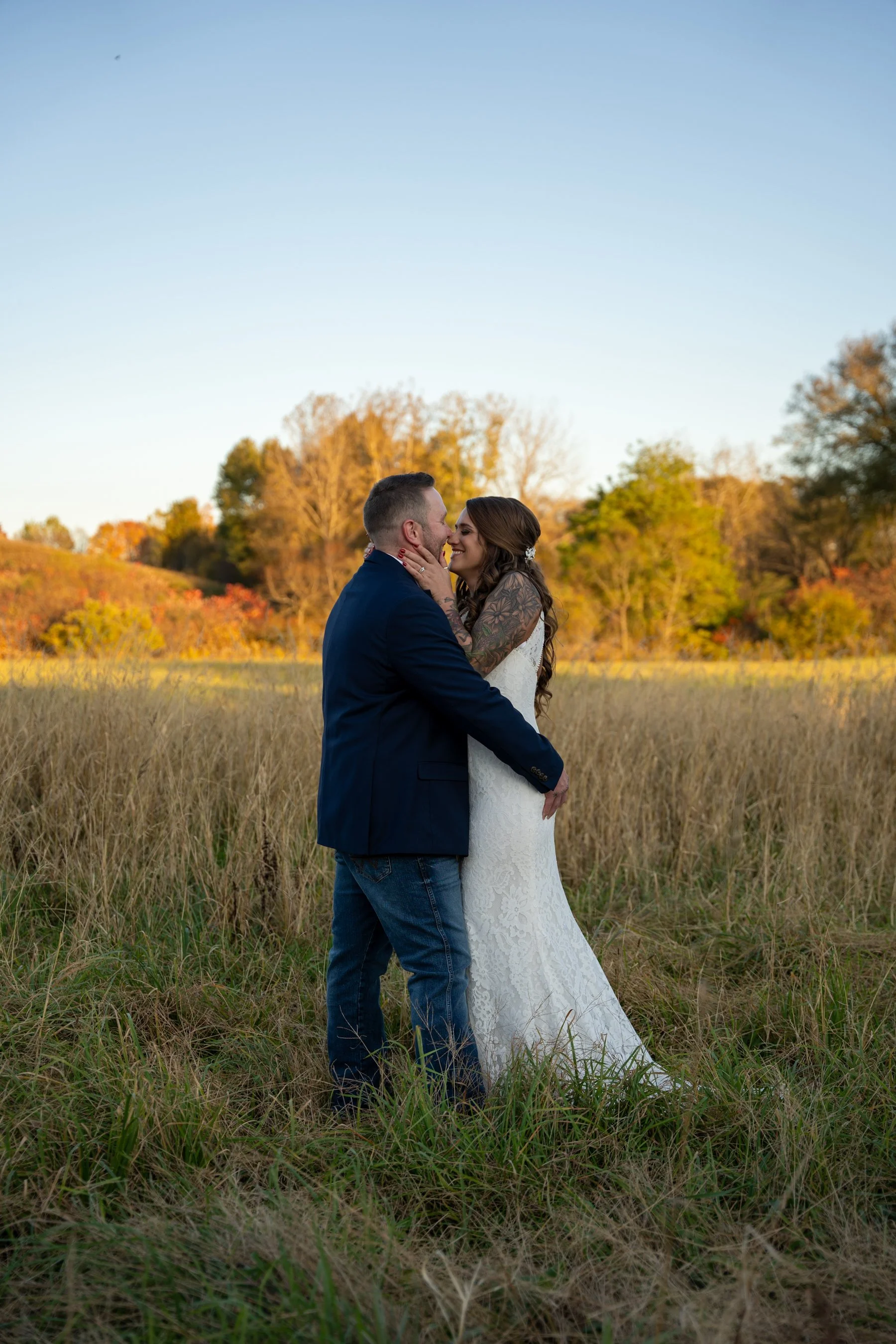 A couple dressed in wedding attire embracing in a field with tall grass, with trees and a clear sky in the background during sunset.