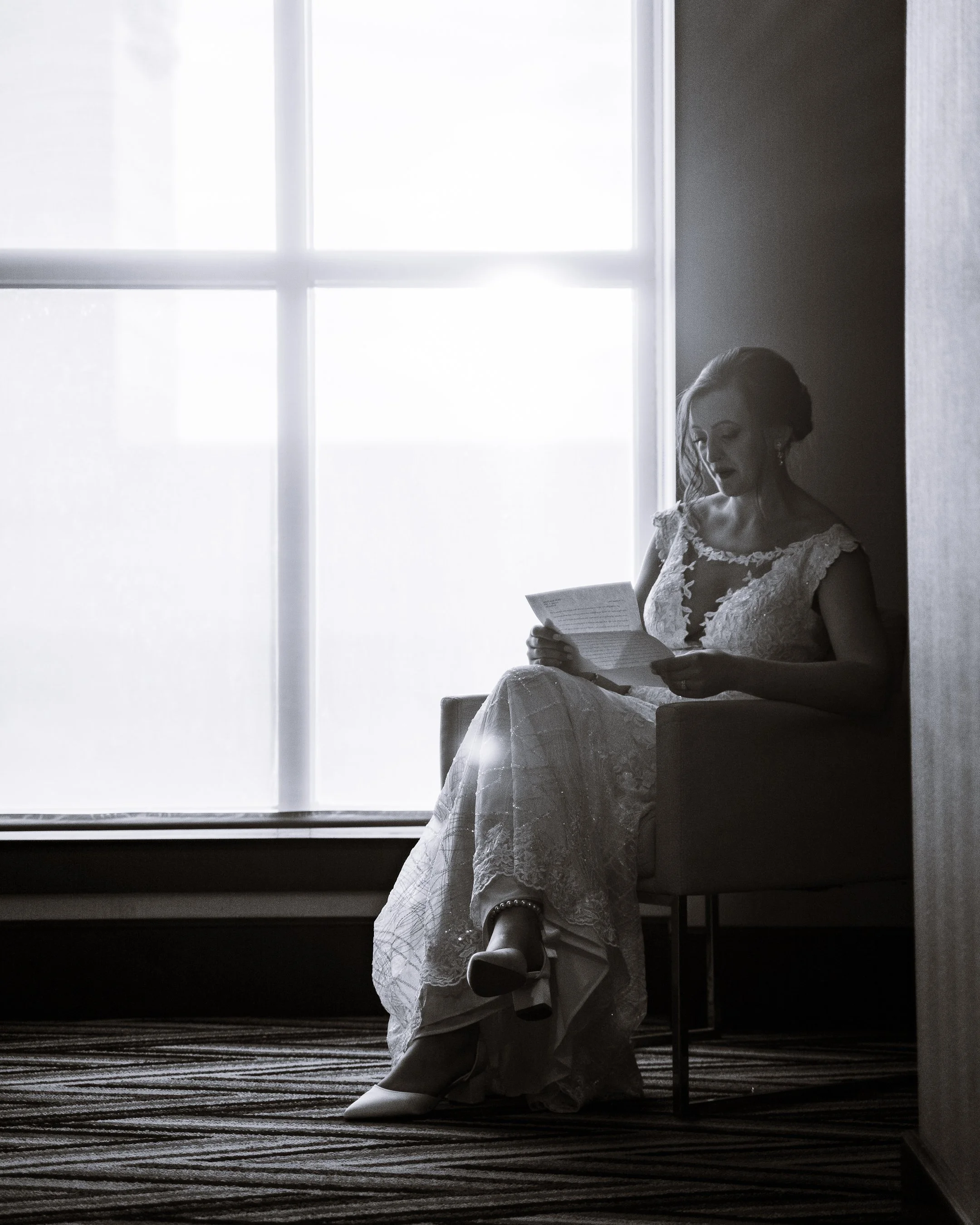 A woman in a wedding dress sitting in a chair by a window, reading a paper.