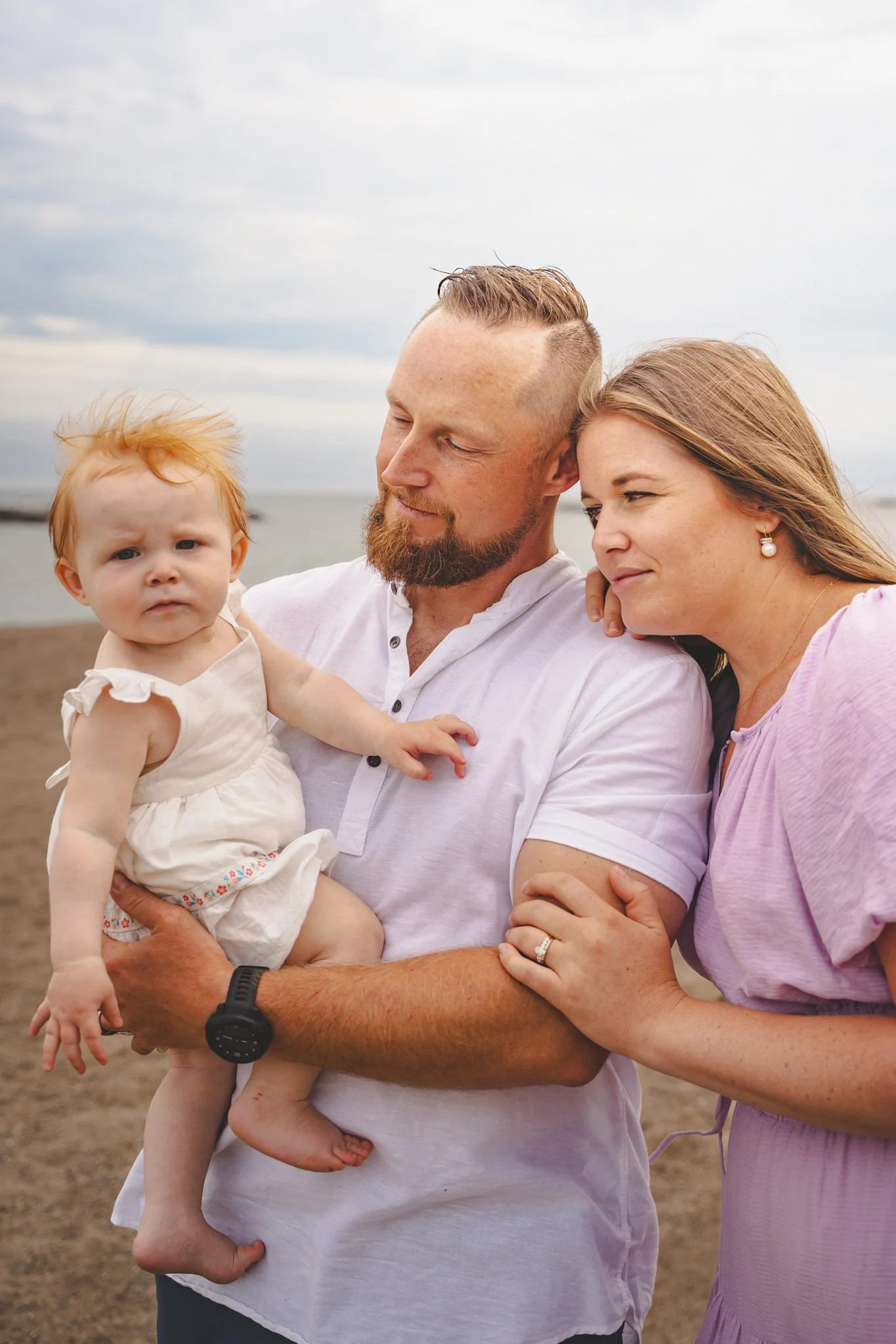 A family of three on the beach during cloudy weather, with the father holding a toddler girl with red hair, while the mother leans on the father's arm.
