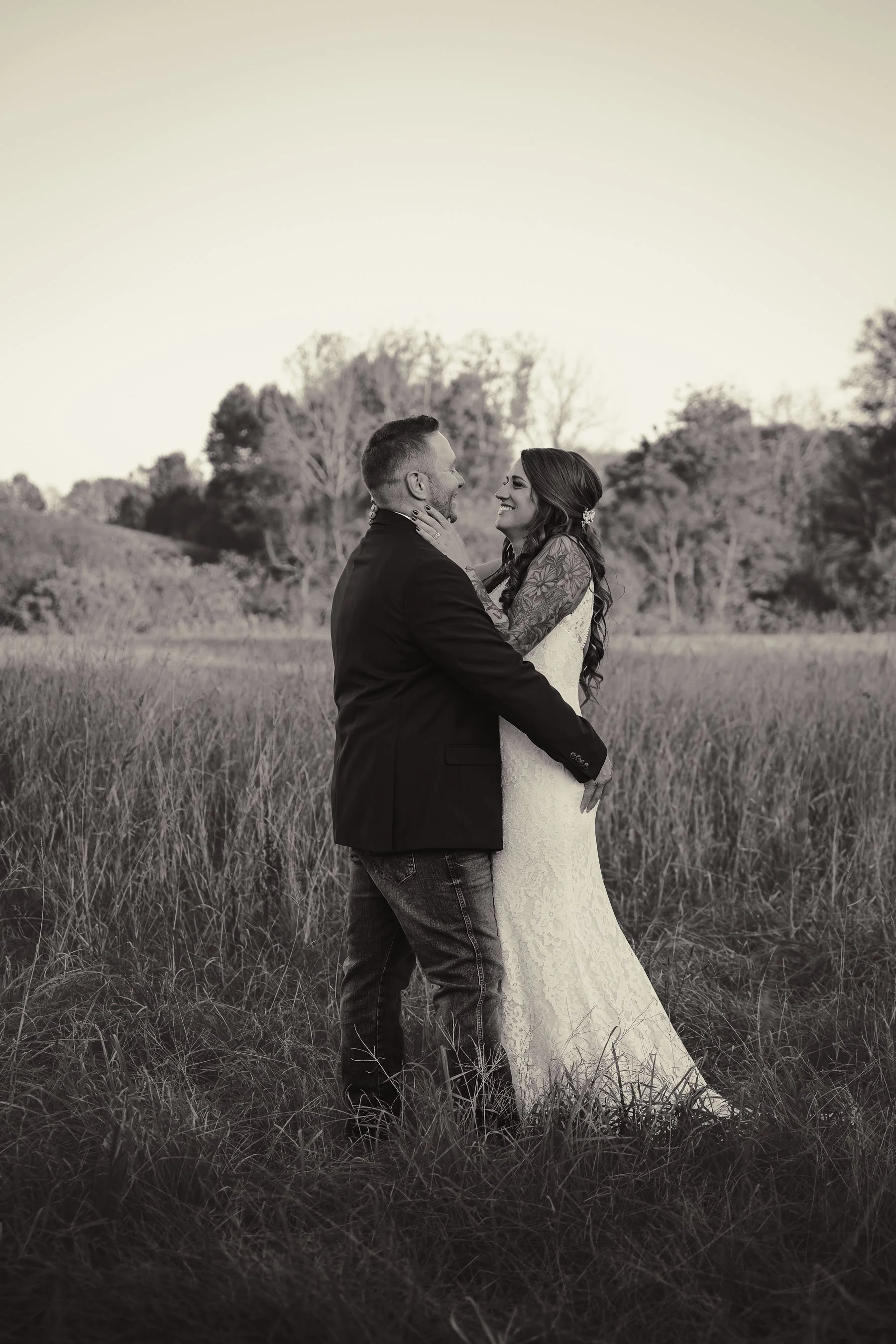 A black-and-white photo of a couple standing in a field, embracing and smiling at each other, with trees in the background.