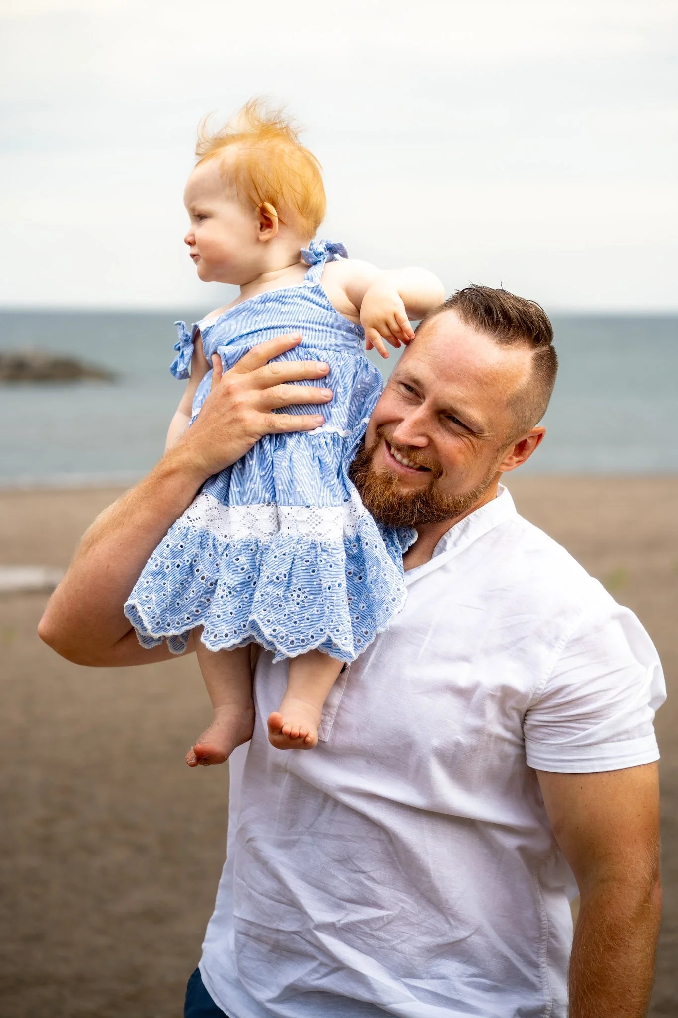 A smiling man holding a young child on his shoulder at the beach with ocean in the background.