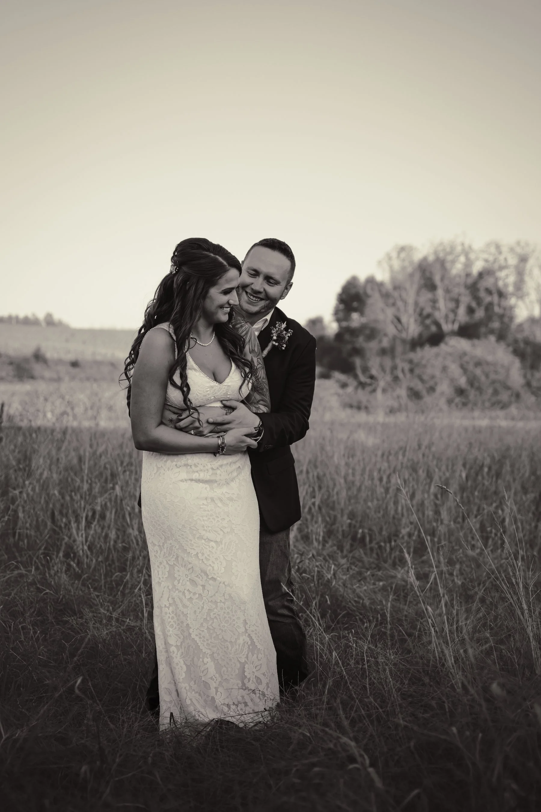 A black-and-white photograph of a couple standing in a grassy field, smiling and embracing each other, with trees in the background.