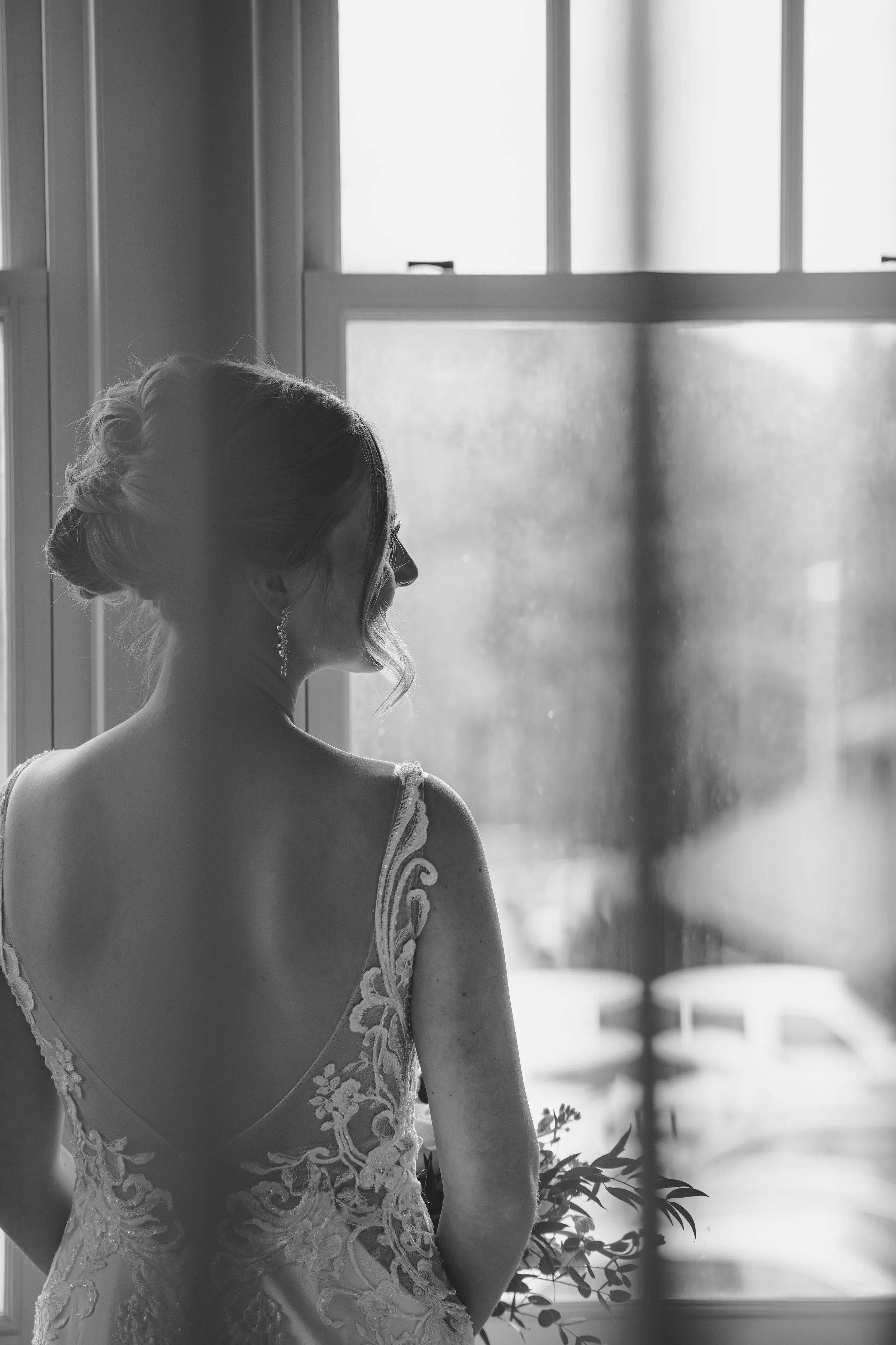 A black and white photo of a woman in a wedding dress looking out a window