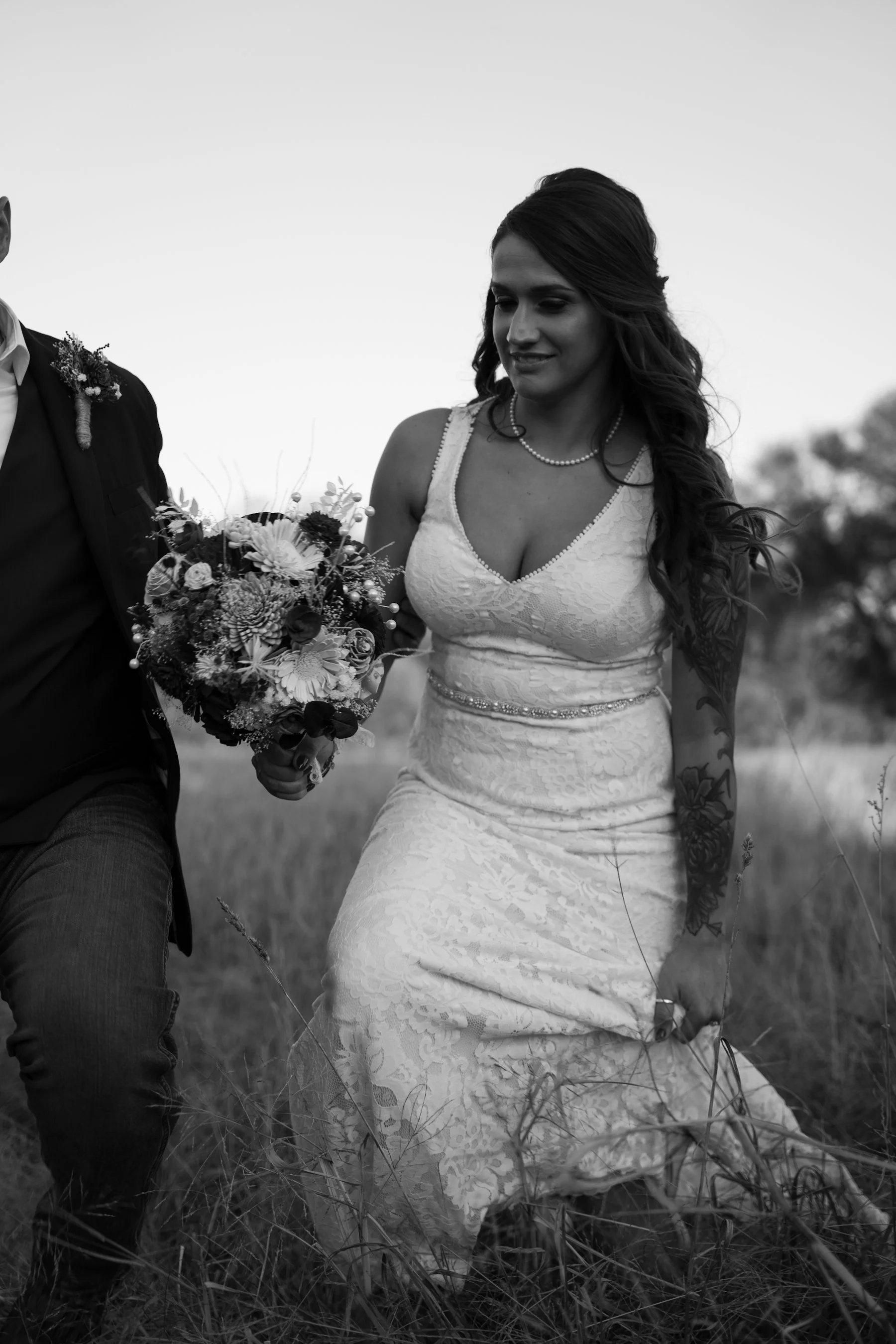 Black and white photo of a woman in a wedding dress holding a bouquet, walking outdoors with a man in a suit partially visible on the left.