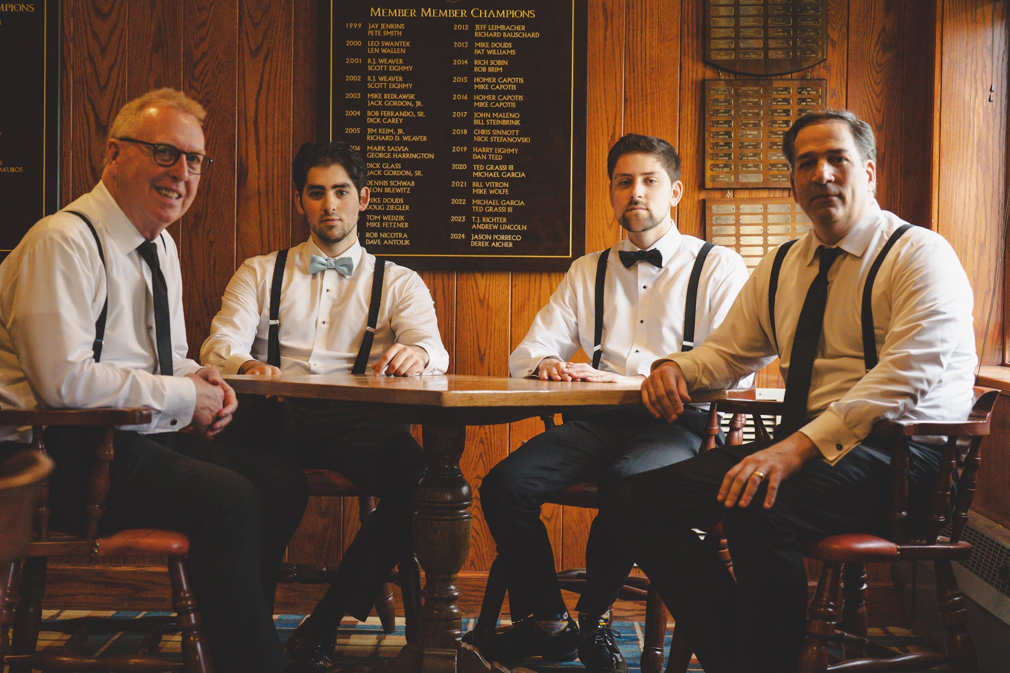 Four men dressed in white shirts, black suspenders, and black pants, sitting at a wooden table in a room with wood-paneled walls and a black plaque listing member champions.