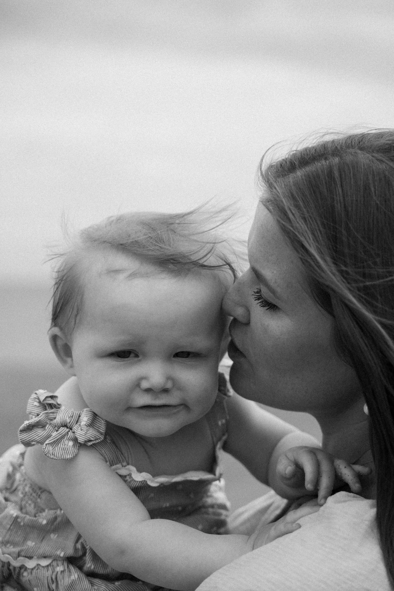 A woman is kissing a young child on the forehead outdoors, with the child's hand resting on her arm. The photo is in black and white.