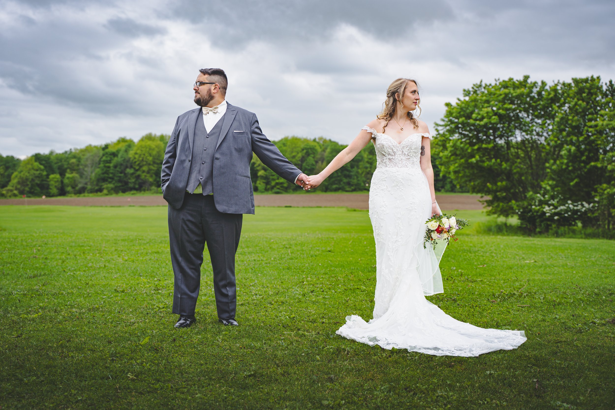 A bride and groom holding hands in a field on a cloudy day, with trees and open land in the background.