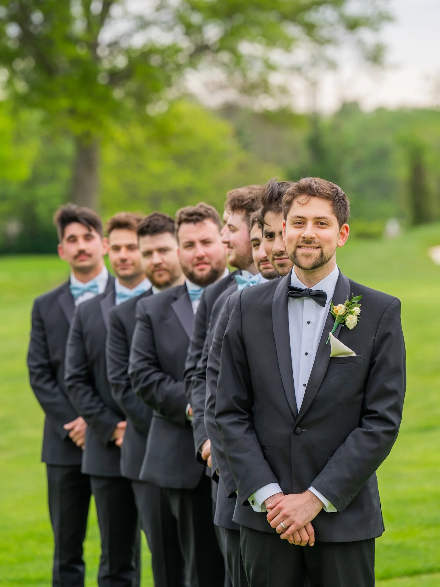 A groom and six groomsmen in formal tuxedos standing outdoors in a line on a grassy field, with trees in the background. The groom, at the front, smiles at the camera.
