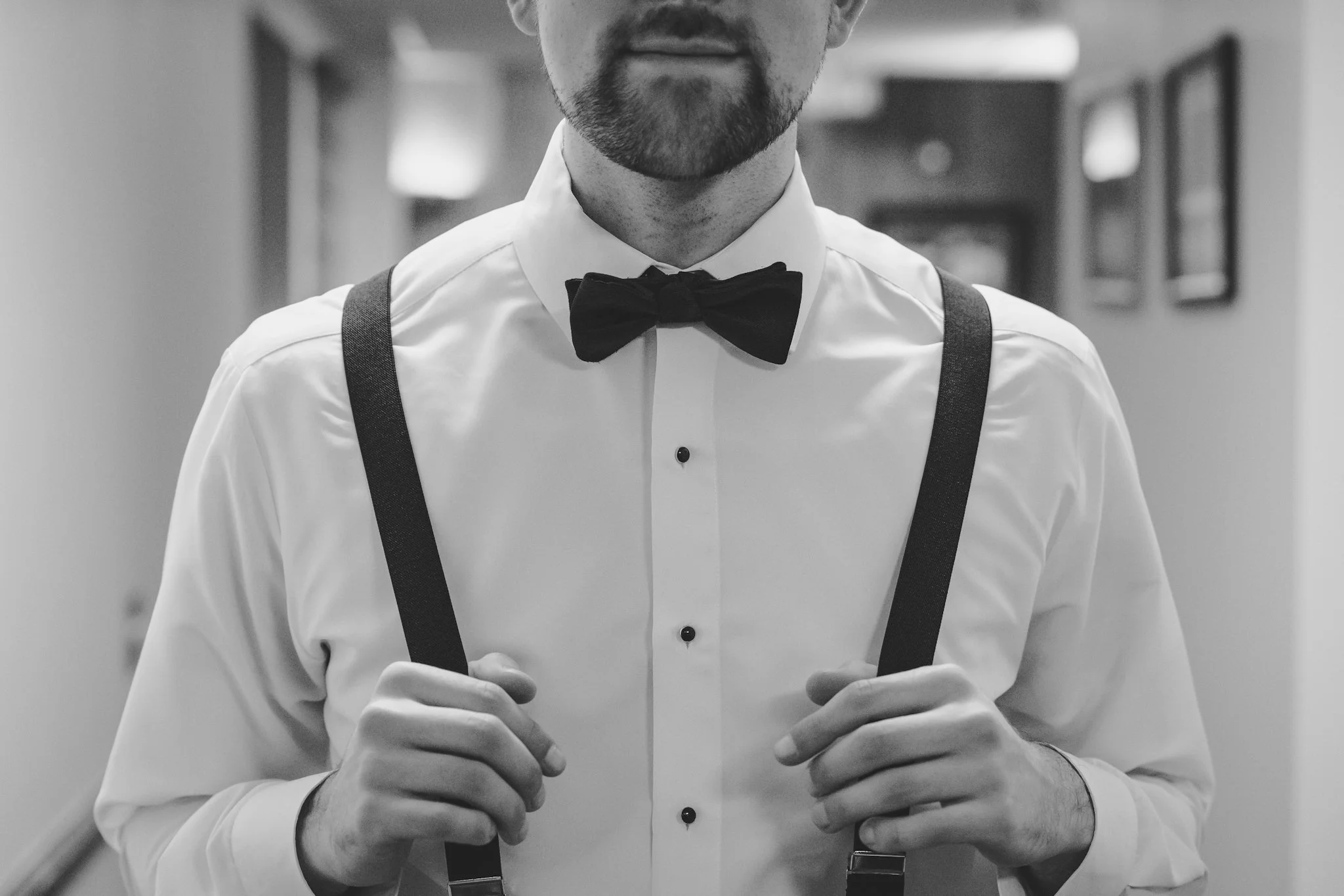 Close-up of a man wearing a tuxedo with suspenders and a bowtie, standing in a hallway with framed pictures on the wall.