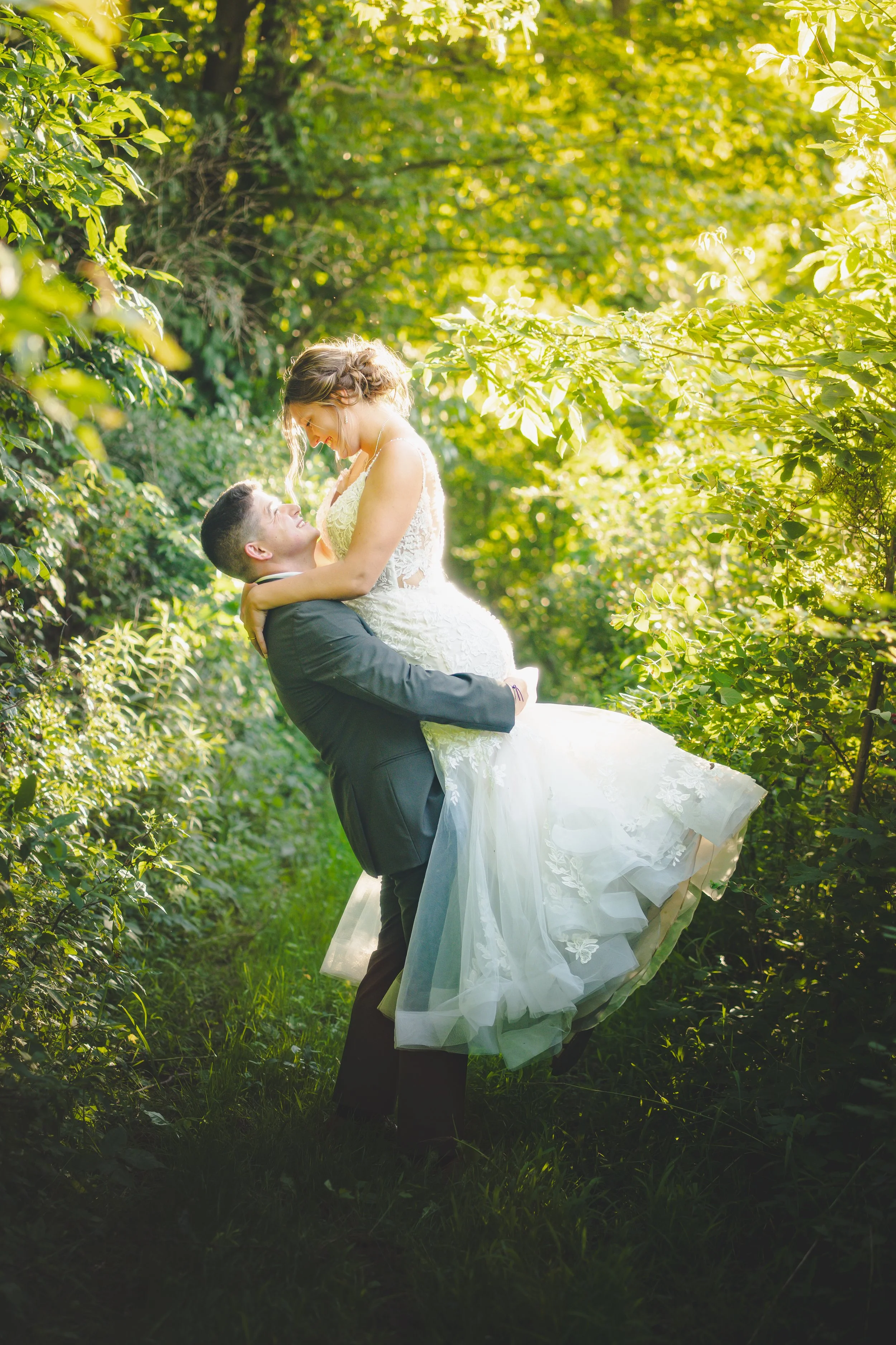 A happy couple in wedding attire during a photo shoot in a green, sunlit forest, with the groom lifting the bride.