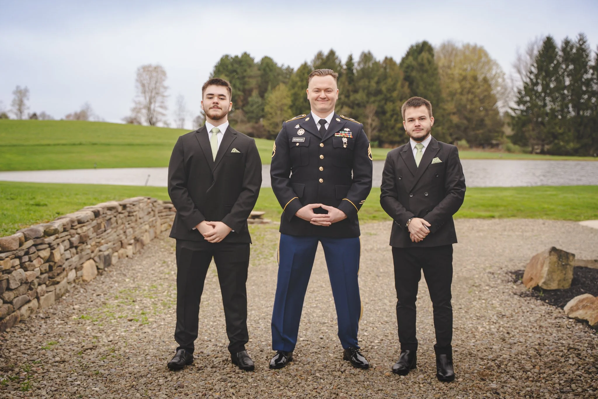 Three men standing outdoors in a park, with a pond and trees in the background. The man in the center is in a military uniform, and the two men on each side are in black suits with light green ties.