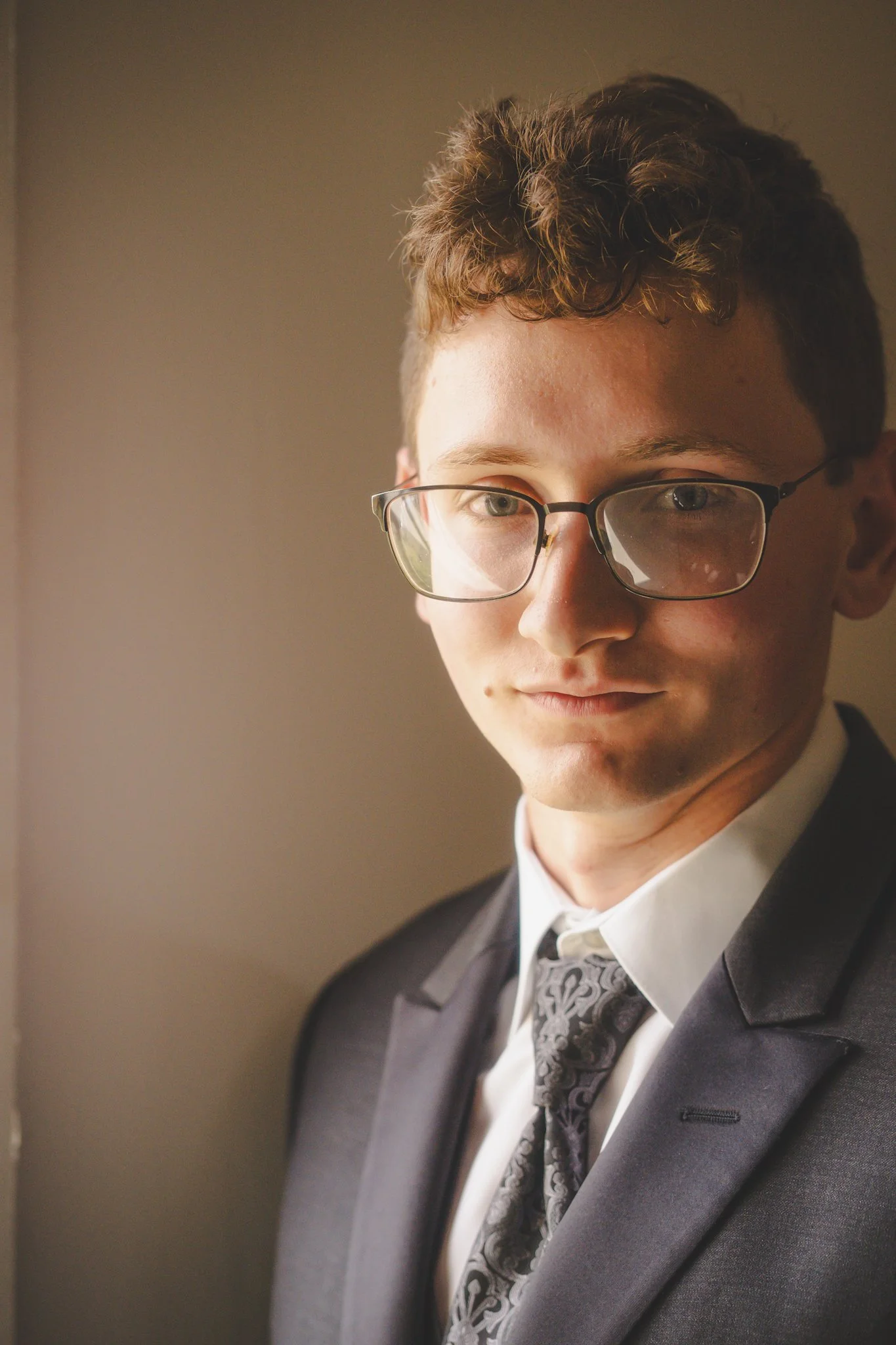 A young man with curly brown hair and glasses is wearing a dark suit, white shirt, and patterned tie, standing by a window with warm lighting.