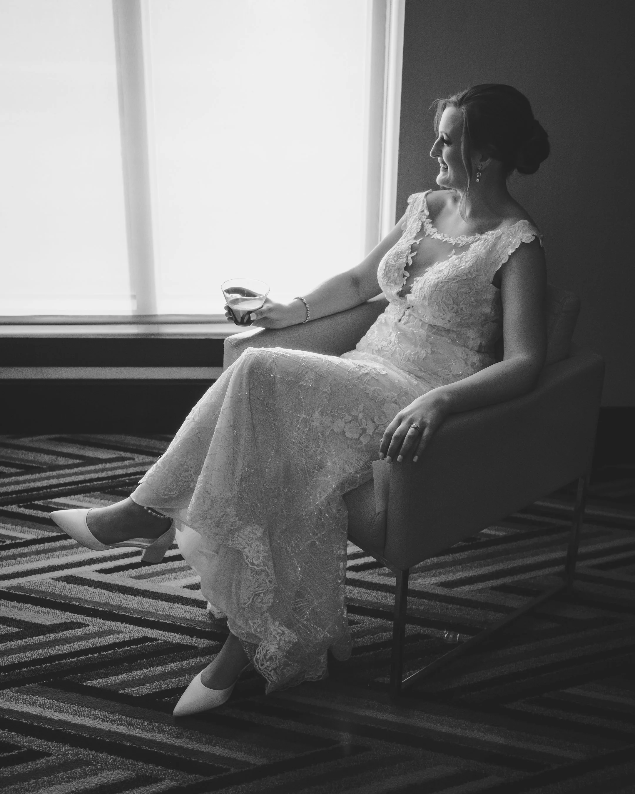 A woman in a lace wedding dress sitting on a chair, holding a glass, smiling, with light coming through a window.