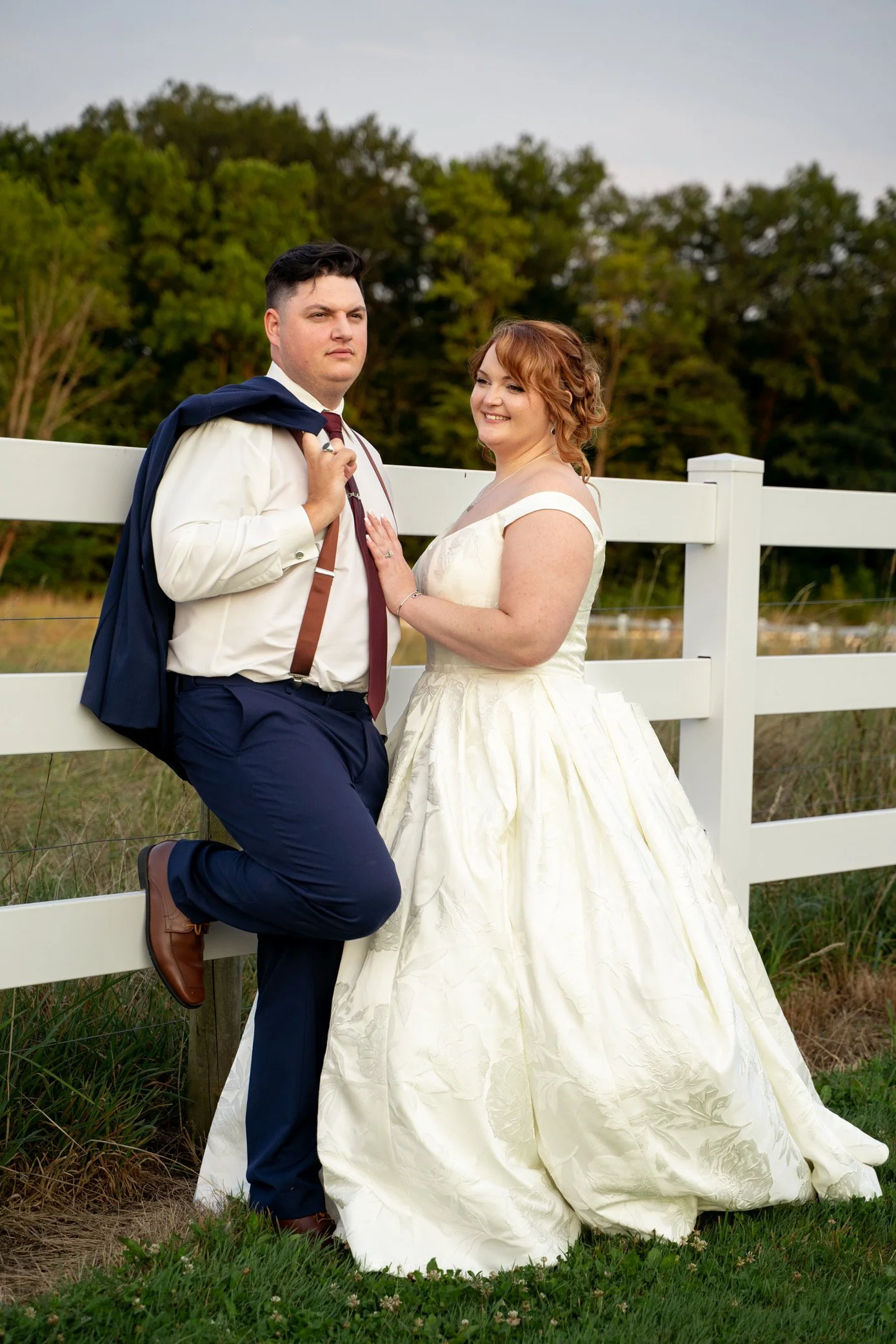A couple dressed in wedding attire standing outdoors beside a white wooden fence, with greenery and trees in the background.