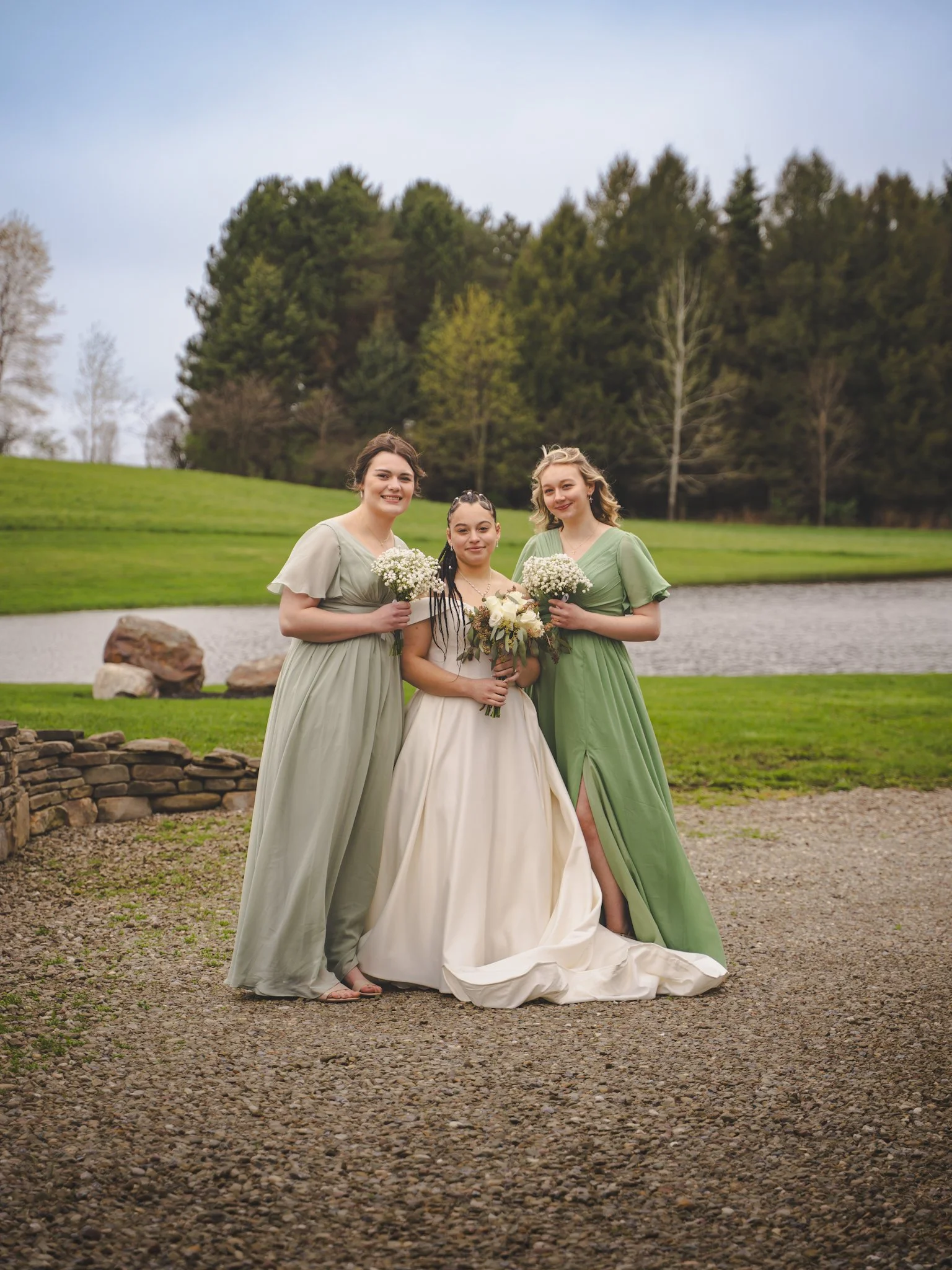 Three women in elegant dresses standing outdoors near a lake and trees, holding bouquets of flowers.