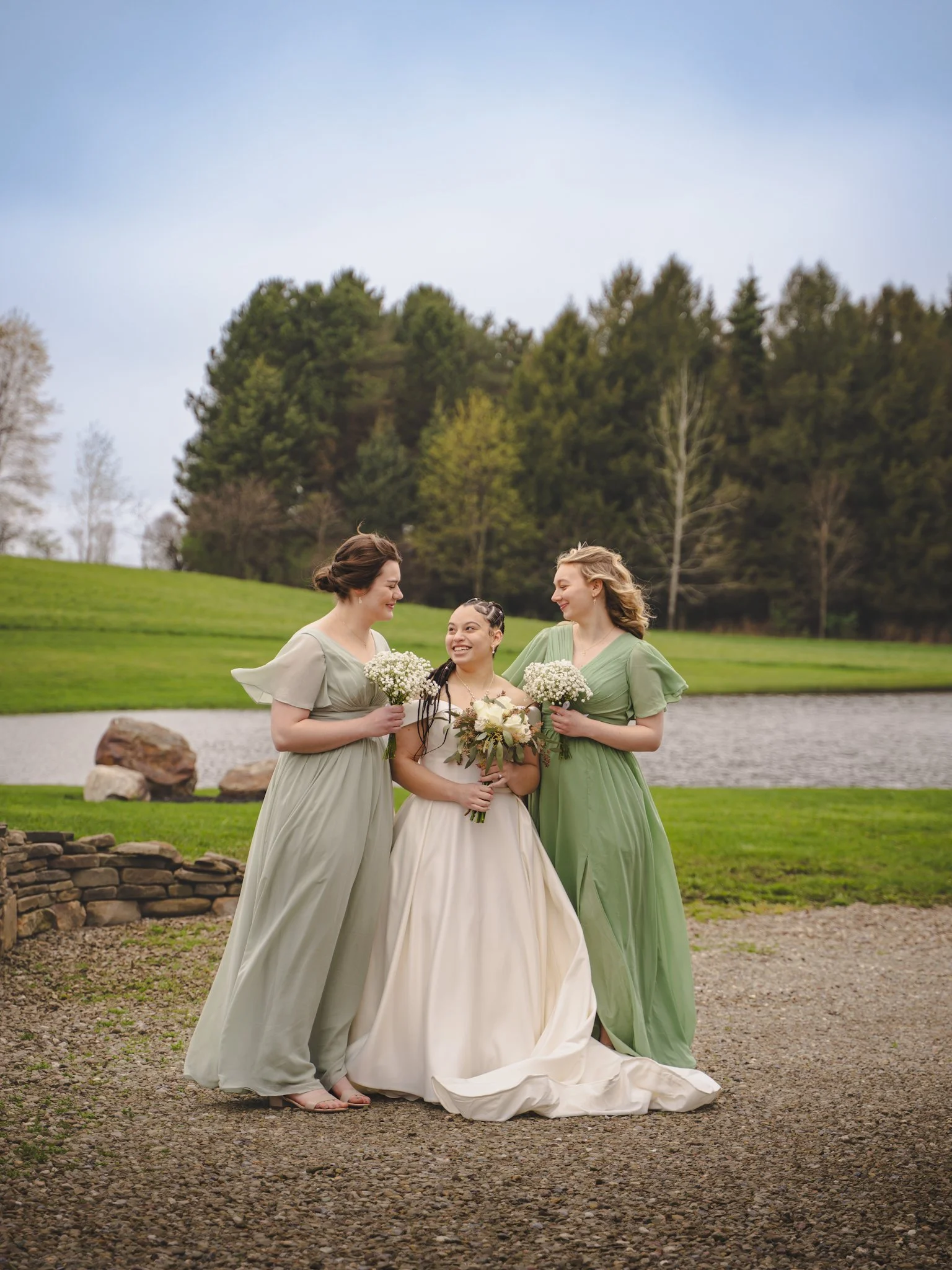 Three women outdoors near a pond, two dressed in green dresses and one in a white wedding gown, holding flowers and smiling at each other.
