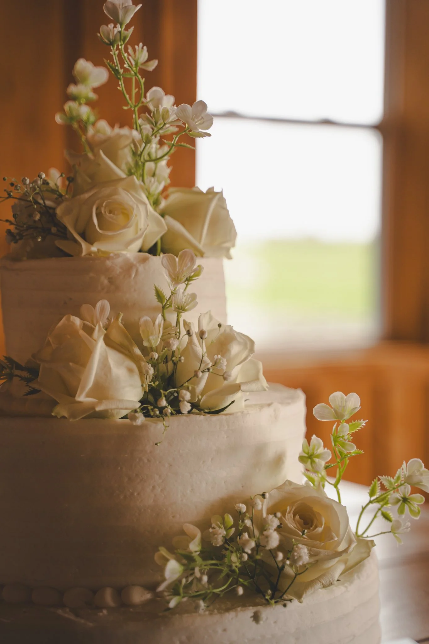 Close-up of a three-tiered white wedding cake decorated with white roses and small white flowers, with a wooden interior and bright window in the background.