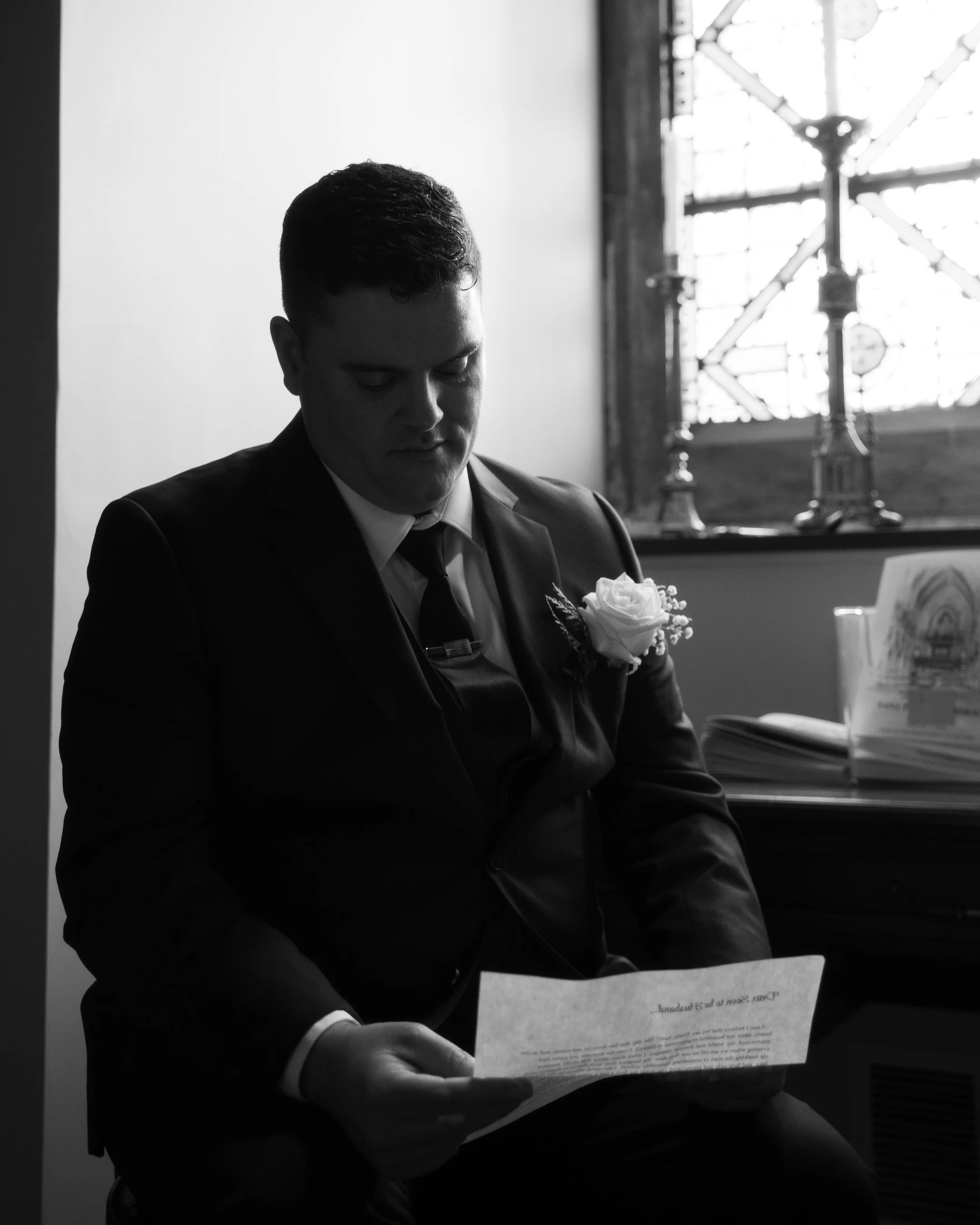 Black and white photo of a man in formal attire reading a piece of paper, with a flower on his lapel, sitting beside a window with stained glass.